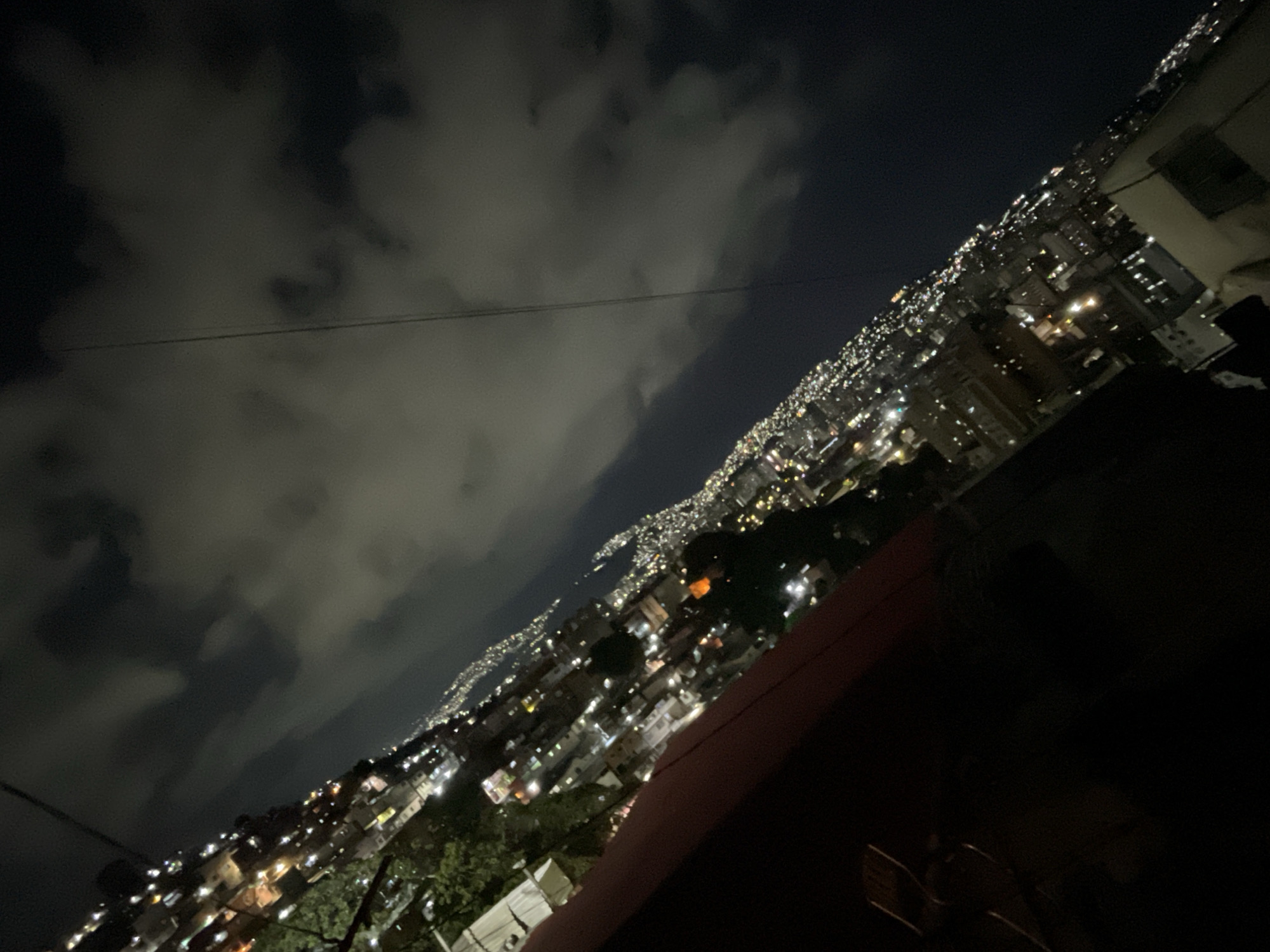 The image depicts a night view of a densely populated area, likely a hillside neighborhood in Caracas, Venezuela. The sky is filled with heavy clouds. Below, numerous lights from houses and buildings illuminate the landscape, stretching into the distance. The lights appear more concentrated toward the center and horizon, suggesting the heart of the city or a more developed area. The foreground is mostly obscured by darkness, featuring a structure that could be a building or a roof. A thin cable stretches across the upper part of the sky.