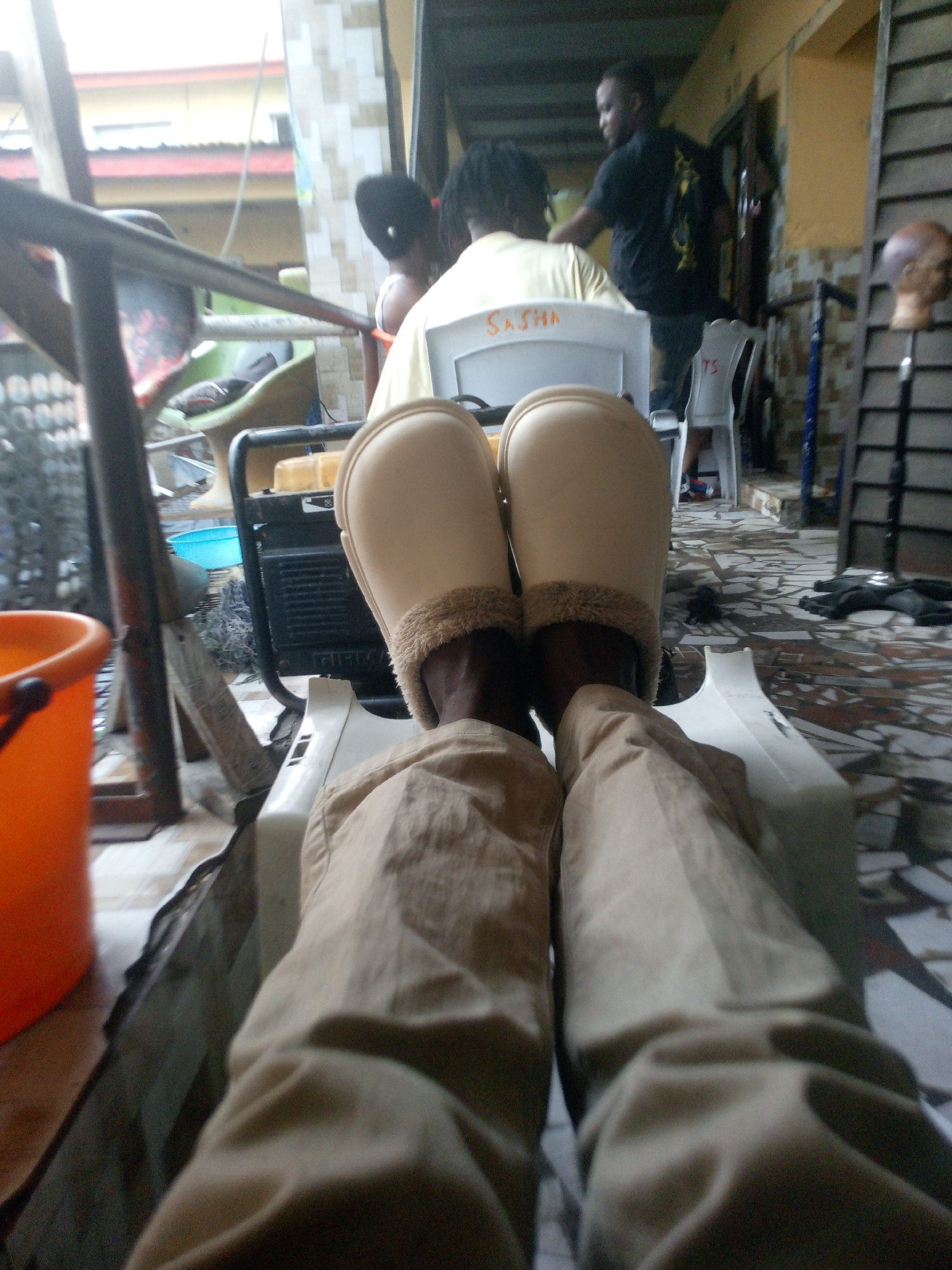 This first-person perspective image captures a casual scene in a semi-outdoor, covered area, possibly a patio or veranda. The viewer's legs, clad in tan/khaki pants, are visible in the foreground, with their feet resting in light-colored, fur-lined slippers.
The floor is covered with patterned grey and white tiles. To the left, an orange bucket is present, and a metal railing runs across the frame. In the mid-ground, a black portable generator is visible, with a blue basin behind it and a yellow object on top.
Several people populate the background. Two individuals are seated in white plastic chairs, facing away from the camera. One chair clearly has "SASHA" written on its back in orange/brown marker, and the person in it wears a light-colored shirt and has dark hair. To their right, a man in a dark t-shirt stands near a doorway. Further to the right, a mannequin head on a stand is visible.
Beyond the covered space, buildings with red roofs and light-colored walls can be seen, indicating a daytime setting, possibly under overcast skies or heavy shade. The scene suggests a relaxed gathering or waiting, with people casually positioned.