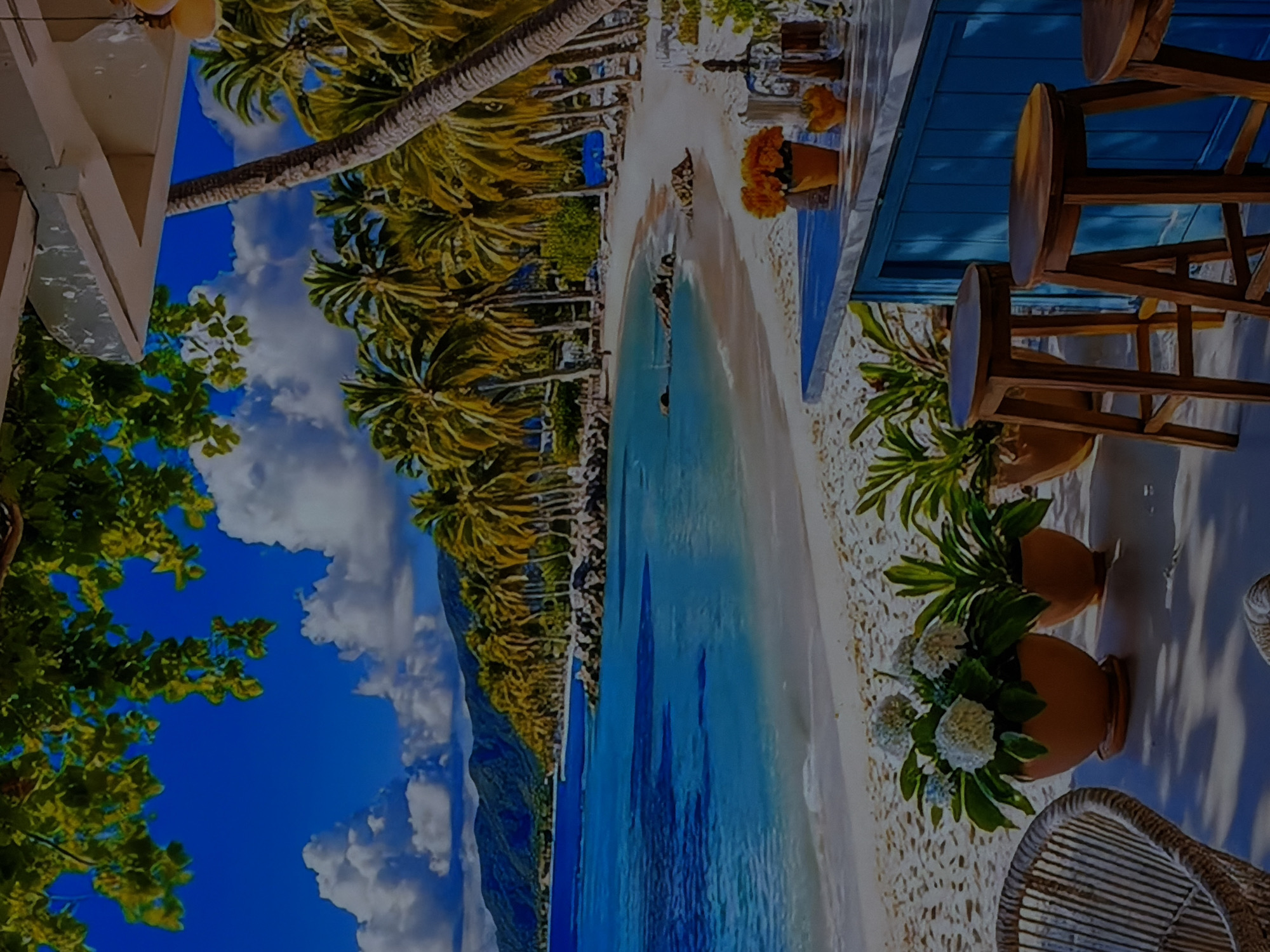 This media file captures a vibrant, sunny tropical beach scene, oriented 90 degrees clockwise from its natural upright position. The foreground on the left shows part of a white building or structure, partially obscured by lush green foliage. A large palm tree trunk extends diagonally across the upper mid-ground, surrounded by dense green palm fronds, suggesting a line of palm trees along the beach.
On the right side of the image, there is an inviting outdoor seating area. It features a bright blue counter or bar, in front of which stand three wooden bar stools with round seats. A small pot of orange flowers rests on the counter. Below the bar, on what appears to be white sand or gravel, are two more potted plants: one with white flowers and another with green foliage. Part of a wicker chair is also visible in the bottom right corner.
The central part of the scene showcases a wide stretch of pristine white sand leading to crystal-clear, turquoise-blue ocean water. The water appears calm and shallow near the shore. In the background, under a bright blue sky adorned with white, fluffy clouds, distant green mountains or hills rise from the horizon.
The overall impression is one of a beautiful, peaceful tropical paradise. The time of day is clearly daytime, with bright sunshine. There are no people visible, suggesting a tranquil atmosphere. No visible text is present in the image. The tropical setting depicted in the image contrasts with the provided location context of Sofiivska Borshchahivka, Ukraine.