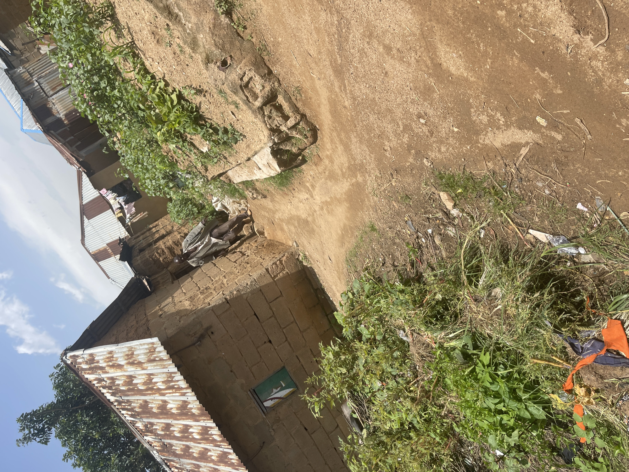 The image depicts a rural or semi-urban street scene in Jos, Nigeria. A man in light-colored clothing, possibly a traditional outfit, is walking down a dirt path. He appears to be of African descent and is facing away from the camera. To the left of the path are buildings constructed from mud bricks and topped with corrugated metal roofs. One building has a small, green-painted window. Lush green vegetation, including some plants that appear to be crops, grows alongside the path and near the buildings. The ground is a reddish-brown dirt. The sky is partly cloudy. The overall impression is one of a simple, possibly impoverished, community.