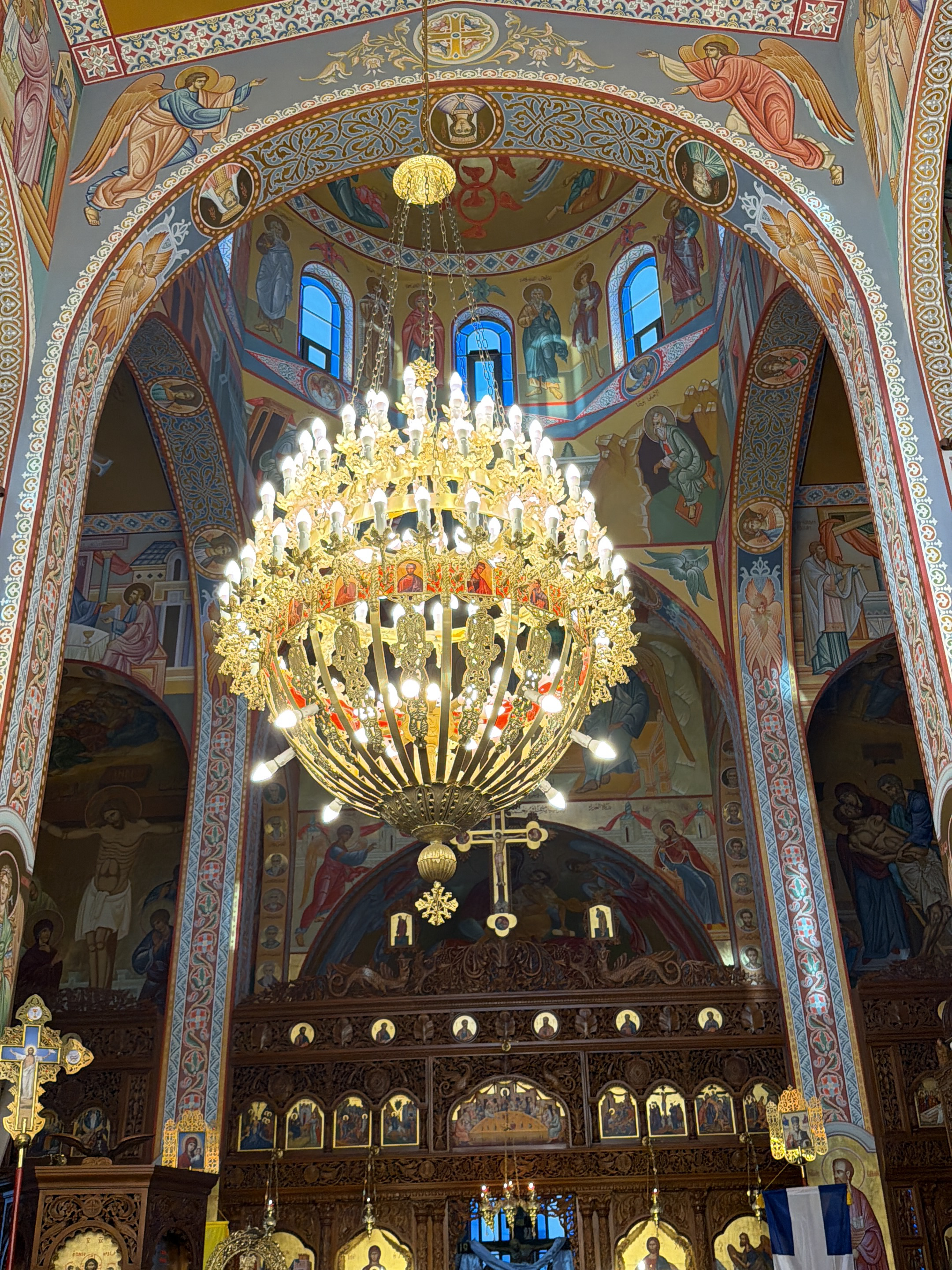 The image captures the richly decorated interior of an Eastern Orthodox church, located in Kafr Yasif, Israel. A massive, multi-tiered golden chandelier, brilliantly lit with numerous bulbs, hangs as the central focal point, illuminating the space.
The walls, arches, and vaulted ceilings of the church are entirely covered with vibrant Byzantine-style murals and icons. These elaborate paintings depict various religious figures and scenes, including angels with outstretched wings, numerous saints, the crucifixion of Jesus Christ, and Mary cradling Christ (Pietà). One prominent mural above the ornate wooden iconostasis depicts the Last Supper. The artistic style features rich colors, predominantly gold, blue, red, and earthy tones, outlined by intricate decorative borders and patterns.
Arched windows, some with blue-tinted glass, are visible higher up, allowing natural light to filter in. The heavily carved dark wooden iconostasis features multiple framed icons of saints and biblical events. Above it, a large golden cross and other golden ornaments are visible. To the left, a golden processional cross with an icon of Christ is present. A blue and white Greek flag is draped on the right side of the iconostasis. Some illegible text in Greek or Arabic script accompanies various figures in the murals. The scene conveys an atmosphere of spiritual grandeur and devotion.