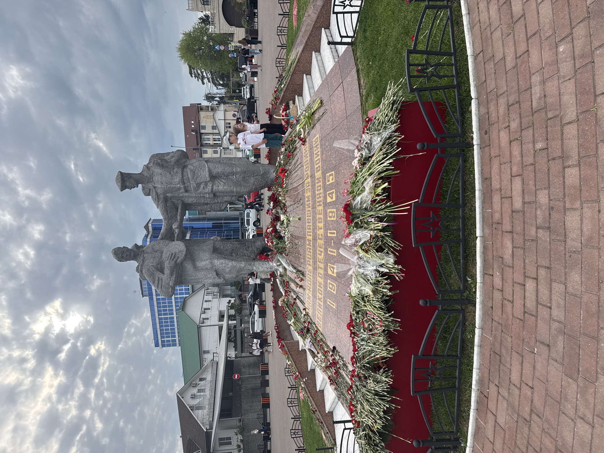The image shows a monument in Sochi, Russia, commemorating the Great Patriotic War (World War II). Two stone sculptures of soldiers stand next to a red and brown stone slab engraved with gold lettering (likely a list of names or a dedication). Numerous bouquets of flowers, primarily red and white carnations, are arranged around the base of the slab. A wrought-iron fence surrounds the area. Several people are standing in the background, including at least two children in white shirts. Various buildings and trees are visible in the background, and the sky is cloudy. The foreground includes a brick walkway.