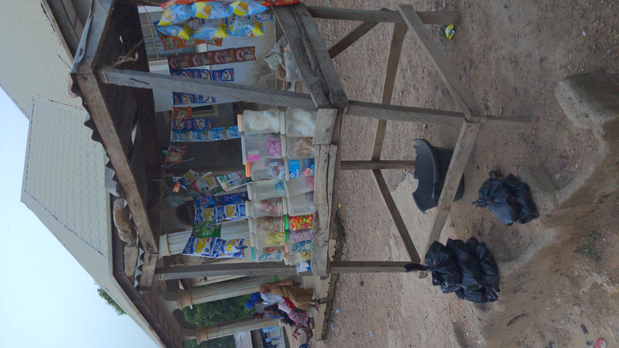 An outdoor market stall or roadside shop is depicted under a clear daytime sky. The stall is constructed from rough-hewn wooden beams and features a corrugated metal roof.
Inside the stall, a variety of products are displayed. Numerous colorful packaged snacks, such as chips and biscuits, hang from strings, forming curtains across the front and interior of the structure. Below these, several rows of clear plastic containers are stacked on wooden shelves, filled with an assortment of loose items including candies and other snacks. The branding on some packages is visible but illegible due to resolution.
In the lower-left quadrant of the image, several individuals are gathered near the stall. These include an adult woman, possibly attending the shop, and multiple children, some partially obscured. They appear engaged in daily activities, potentially shopping or socializing.
The ground surrounding the stall is an unpaved dirt or sandy surface. To the right of the wooden structure, a black plastic basin or bucket is visible, alongside several black plastic trash bags resting on the ground. A light-colored building with a patterned roof is partially visible in the upper-left background. The scene portrays a bustling, informal retail environment.