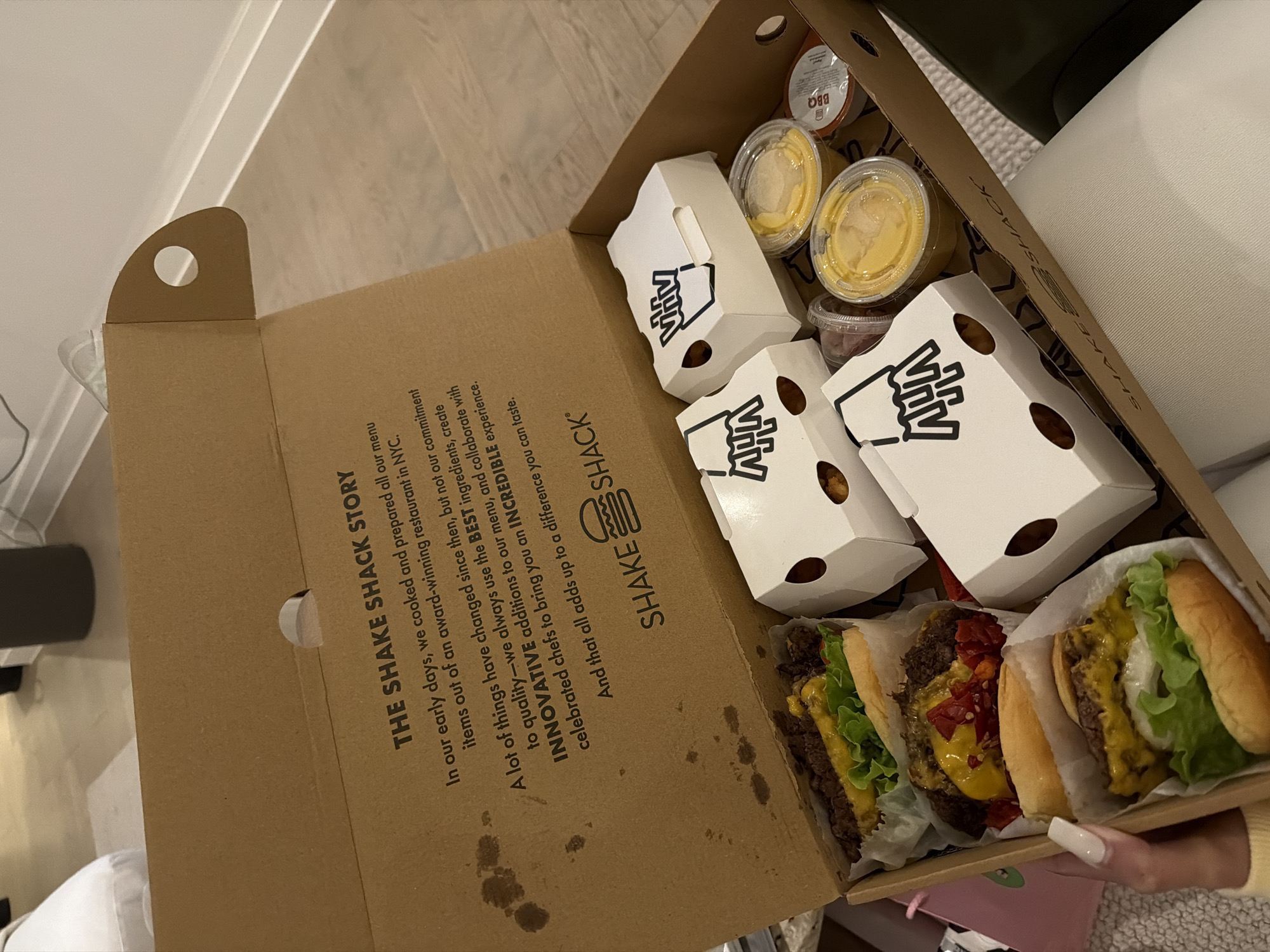 A close-up, high-angle shot captures an open Shake Shack takeaway box, revealing a meal intended for consumption in an indoor setting, likely a home in London, United Kingdom. The box contains three cheeseburgers, two of which appear to be double patties, with one featuring visible red peppers or relish. Alongside the burgers are three white cardboard containers with a stylized fries logo ("Mhiv"), likely holding French fries or tater tots. Several small plastic sauce cups are also present; two contain a yellow, cheese-like sauce, another holds a reddish sauce, and one is clearly labeled "BBQ". A person's hand with long, light-colored nails is visible at the bottom right, gently holding or supporting the box.
The interior of the cardboard box lid features text detailing "THE SHAKE SHACK STORY," which describes their commitment to using "BEST ingredients," creating "INNOVATIVE additions," and collaborating with "celebrated chefs" to deliver an "INCREDIBLE experience" that one "can taste." Below this text is the Shake Shack logo, depicting a stylized burger. The background shows parts of a light-colored wooden floor and white upholstered furniture, suggesting a casual, domestic environment. The lighting is bright and even, indicative of daytime or a well-lit interior. The overall impression is one of a convenient and enjoyable fast-food meal about to be consumed.