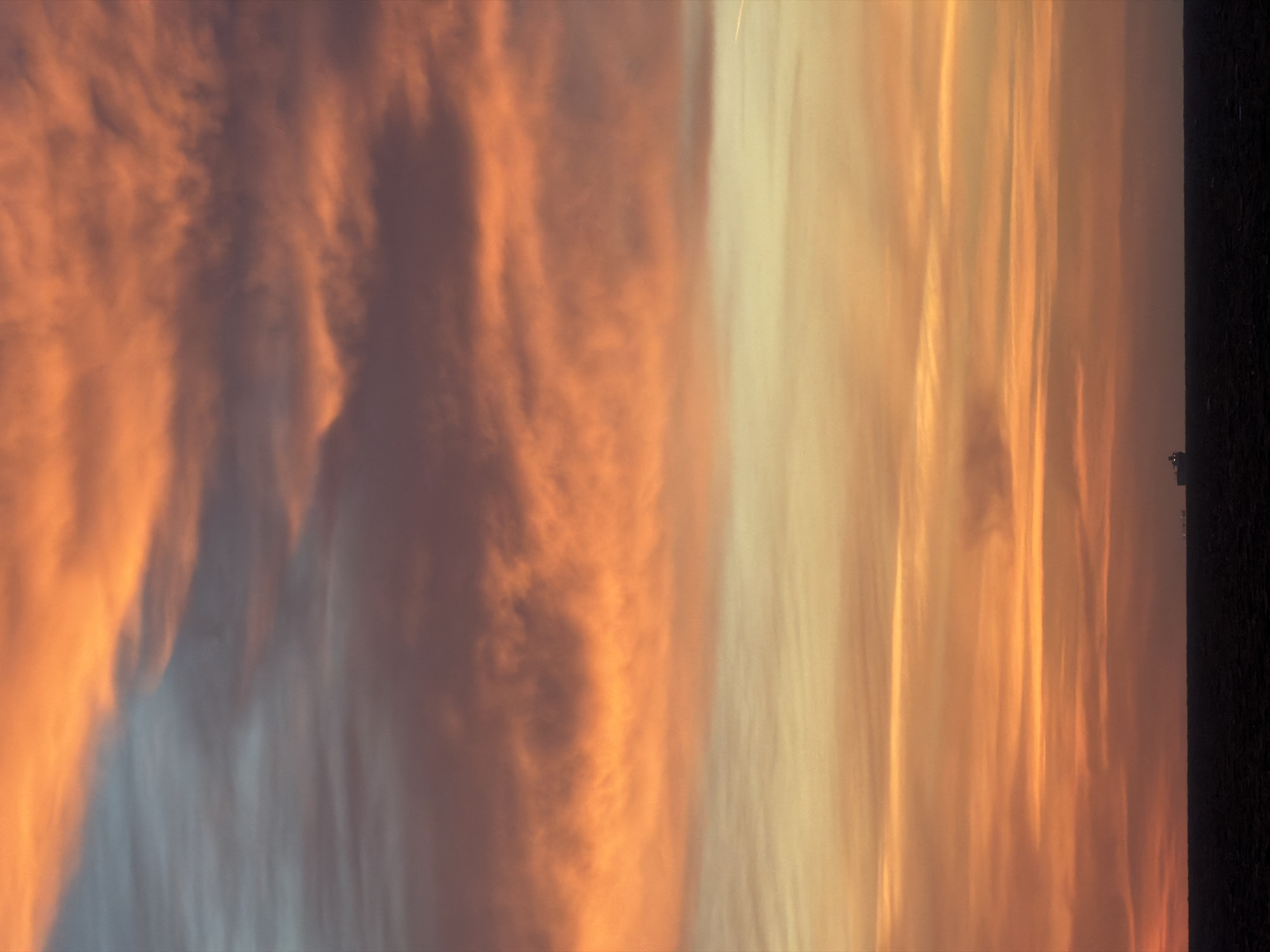 The media file displays a sky scene dominated by dramatic cloud formations, photographed during either sunrise or sunset. The image appears to be rotated 90 degrees clockwise. When viewed in standard landscape orientation, the low-angle light source seems to be coming from the right, casting warm hues across the clouds.
The left side of the sky is filled with voluminous, textured clouds displaying a rich palette of deep oranges, reddish-browns, and purplish-grey shadows, creating a sense of depth and movement. Towards the right side, the clouds transition into more elongated, streaky formations, bathed in softer shades of yellow, cream, and pale orange, indicating direct illumination by the setting or rising sun. A very thin, bright, vertical line, possibly a distant airplane contrail or a highly illuminated wispy cloud, is faintly visible in the upper right quadrant of the image.
On the far right edge of the image, a dark, vertical strip occupies a small portion of the frame. This indistinct feature could represent a distant landmass, a building, or part of the immediate foreground, but its details are too obscured for positive identification.
There are no discernible human subjects or specific activities taking place; the image captures a natural phenomenon. The warm and vibrant colors evoke a serene and majestic atmosphere, characteristic of twilight hours. No visible text is present in the image.