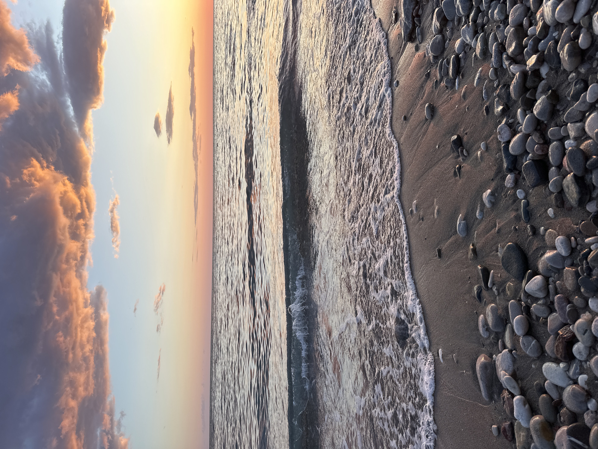 This image captures a serene coastal scene at either sunrise or sunset, set in Šušanj, Montenegro. The frame is horizontally divided into distinct elements: sky, sea, and shoreline.
The upper portion of the image is dominated by a vibrant sky, transitioning from a soft blue on the left to warm yellow and orange hues near the horizon on the right, indicative of golden hour. Several clouds, illuminated by the low sun, display rich orange and pink tones, particularly a large, voluminous cloud cluster on the far left.
Below the sky lies the calm sea, its surface reflecting the warm colors of the sky. Gentle ripples stretch across the water, catching the light. A wave is breaking or receding onto the shore, forming a line of white foam that marks the boundary between the water and the land.
The foreground features the pebbly and sandy beach. Numerous smooth, grey and dark pebbles of various sizes are scattered across the sand, extending from the water's edge up towards the viewer. The wet sand near the receding wave also shows reflections of the soft light.
There are no people or man-made objects visible, emphasizing the natural beauty and tranquility of the scene. The overall mood is peaceful and calm. There is no visible text in the image.