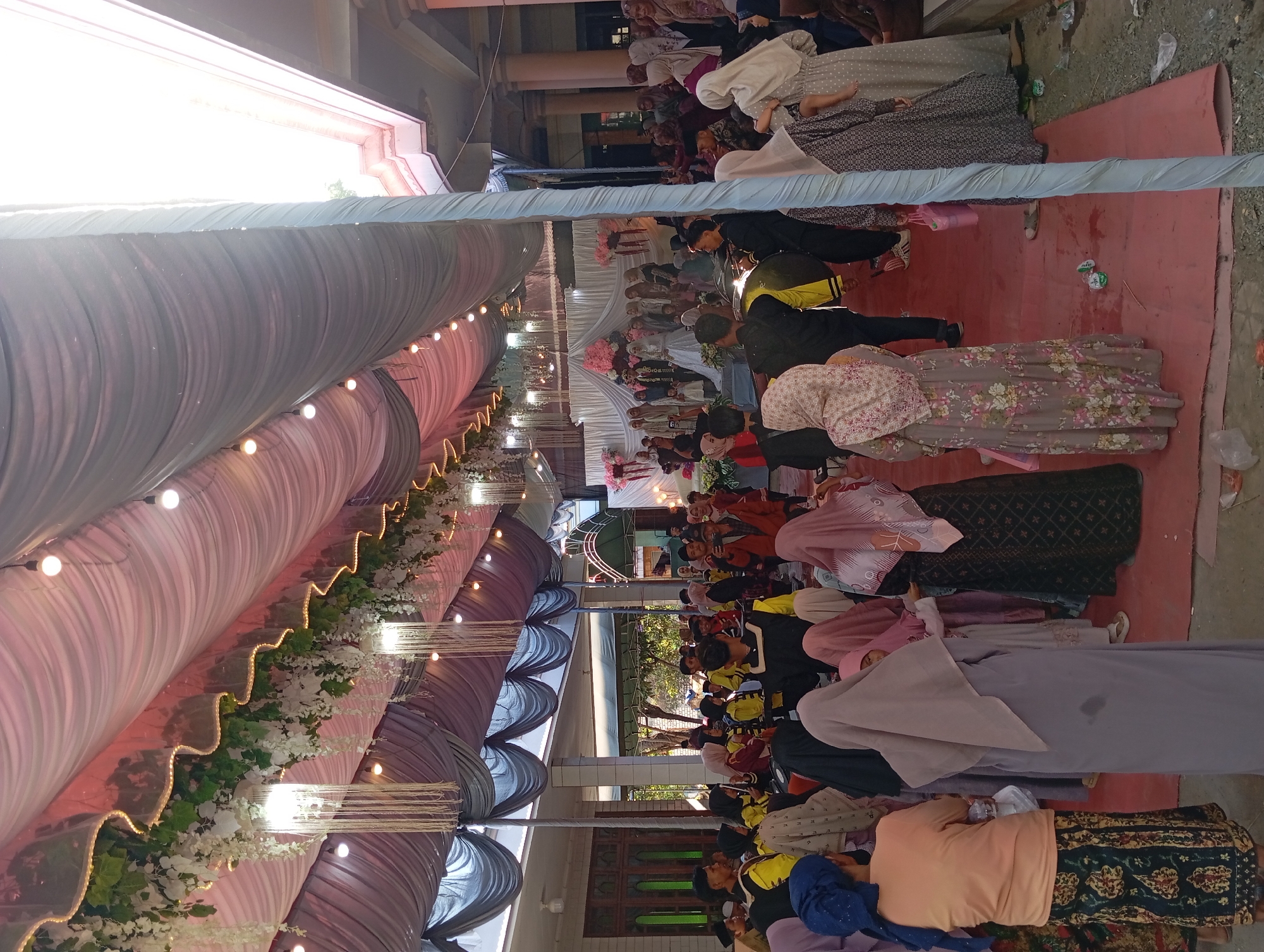 This is a candid, medium-shot, outdoor photograph taken during a wedding ceremony in Padurungan, Indonesia. The scene is under a large, draped tent with pink and gray fabric. White floral garlands and string lights are suspended from the tent ceiling. A red carpet covers the ground. In the background, a bride in a white gown and a groom in a dark suit stand on a raised platform, surrounded by guests. The foreground features a gathering of people, many of whom are wearing Islamic attire, including hijabs and modest dresses. Some individuals are holding cameras, suggesting they are documenting the event. The atmosphere appears festive and communal, with guests mingling and observing the ceremony. The environment suggests a warm, possibly humid, climate common in Indonesia.