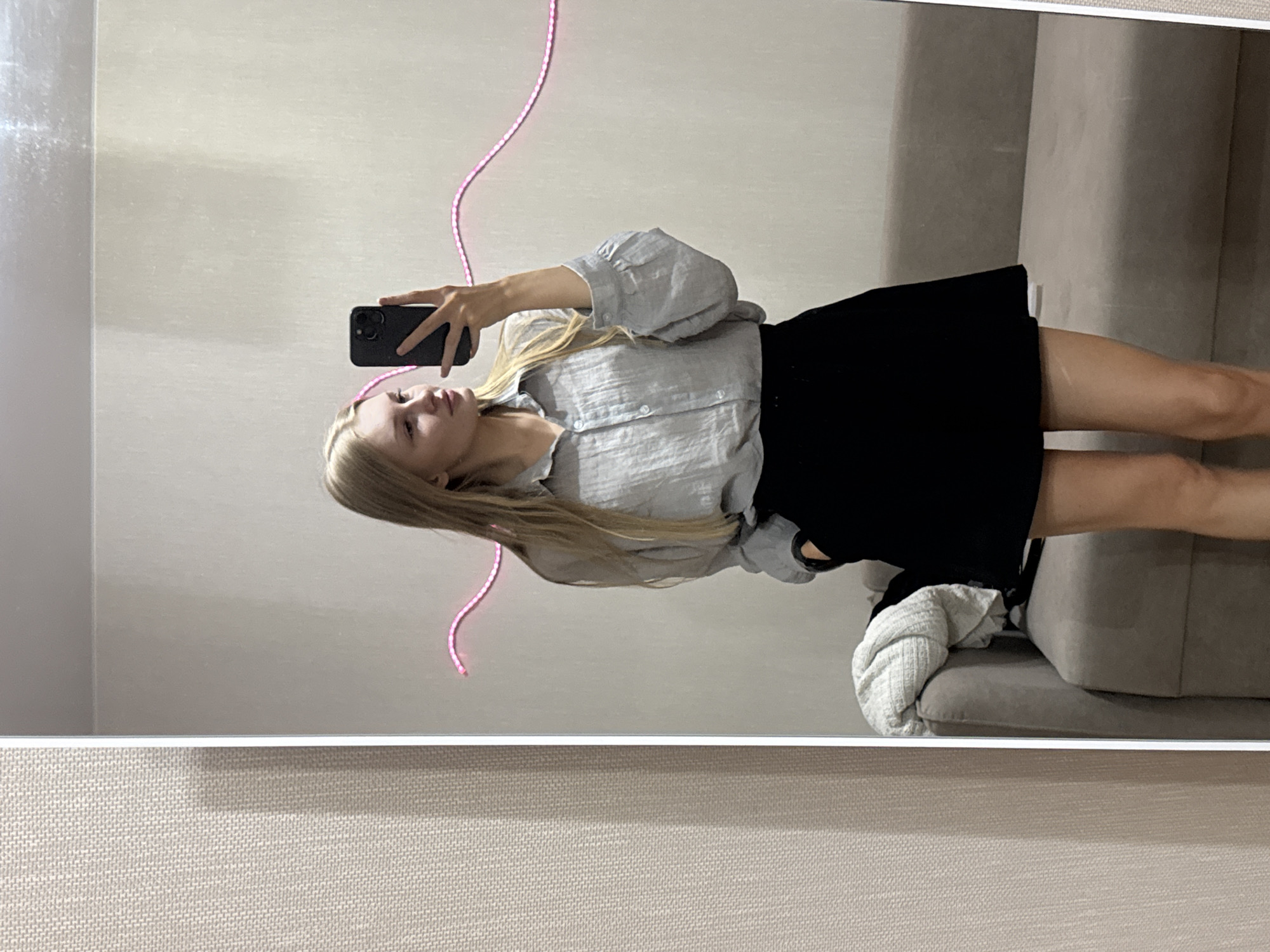 A young woman with blonde hair is taking a mirror selfie in a room. She is wearing a light grey, oversized, button-up shirt and a black skirt. Her left hand is holding her phone, and her right hand is in her pocket. A pink neon light strip is visible on the wall behind her, shaped like a wave. To the left of the woman, a grey sofa with a white blanket draped over it is partially visible. The room appears to be a bedroom or dressing area, with a textured wall to the left of the mirror. The overall lighting is soft and natural. The image does not provide any specific landmarks or clues to pinpoint the location within the city of Perm, Russia.