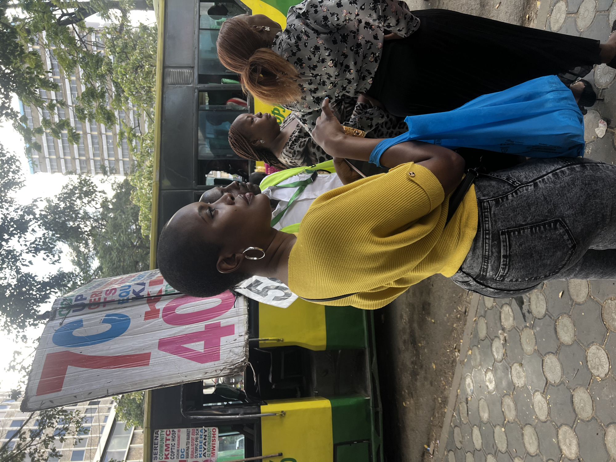The image shows a group of people in Nairobi, Kenya, next to a matatu, a shared taxi. The matatu is painted in yellow and green, with a sign displaying "72, 40" and a list of destinations, including "Kibera, Ayany, Mwisho". A woman in a yellow sweater and grey jeans is in the foreground, looking up and to the left. Behind her is another woman with reddish-brown hair, wearing a black dress and floral blouse. The presence of these individuals suggests they are waiting for or interacting with the matatu, likely as passengers or in transit. The background includes a building and trees, indicating the setting is urban. The presence of a matatu and the associated text on the vehicle indicate that the location is Nairobi.