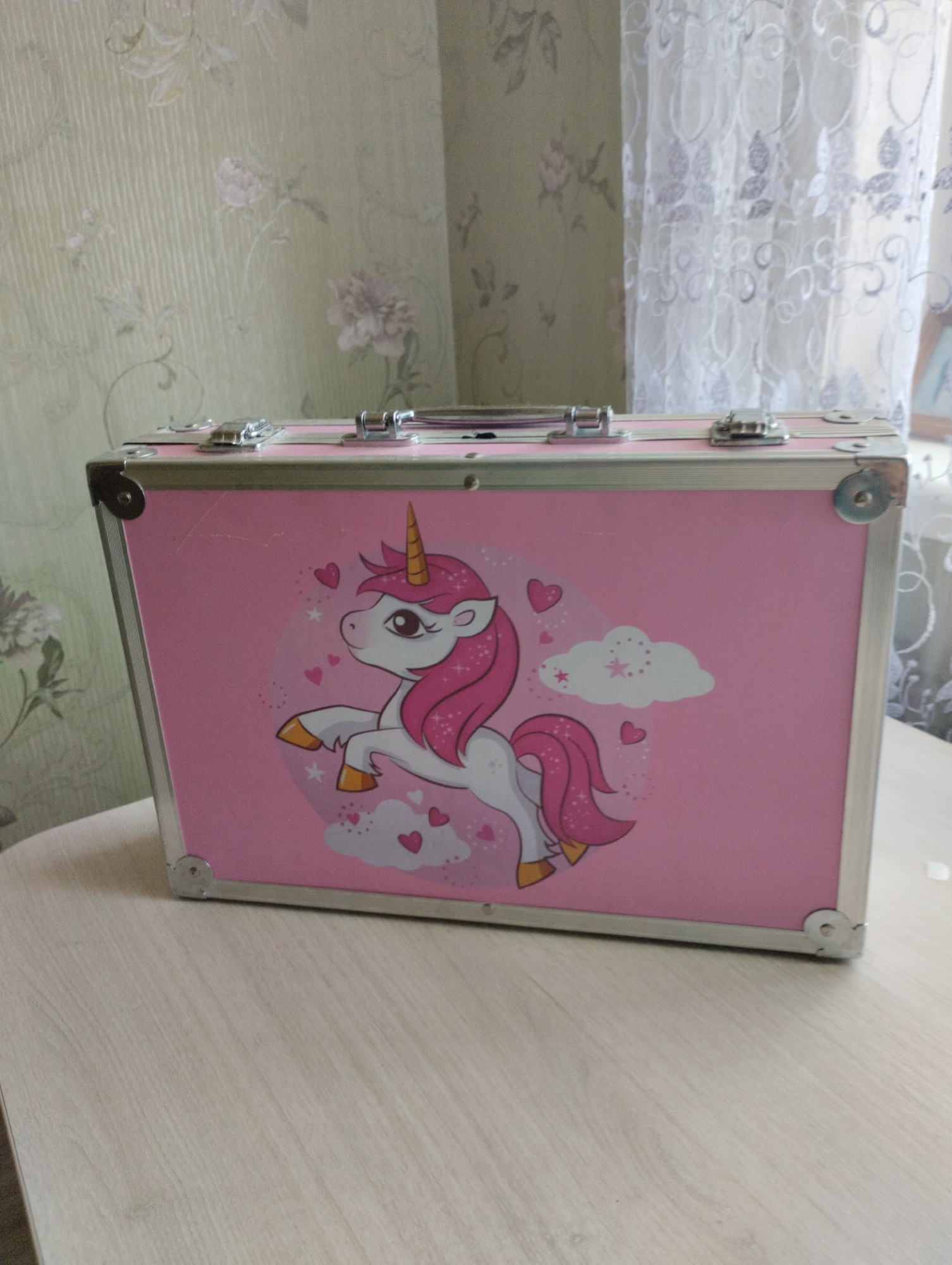 A pink, rectangular, hard-sided case with silver trim and clasps is displayed prominently on a light-colored wooden table. The case features a colorful illustration of a unicorn with a pink mane and tail, surrounded by hearts and clouds. The background consists of a wall with floral wallpaper and a sheer white curtain. The scene is set indoors. The image is taken from a frontal, slightly elevated perspective. No people are visible. The overall impression is one of a child's item, possibly a makeup or toy case. The location is Astravyets, Belarus.