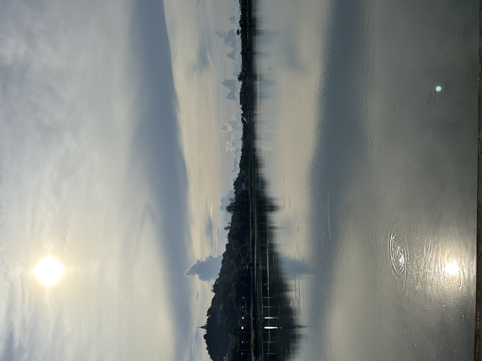 The image shows a body of water, likely a lake, reflecting the sky and surrounding landscape. The sky is partly cloudy, with the sun visible and positioned on the left side. On the opposite bank of the water, there's a tree-covered hill with buildings and streetlights. Ripples disturb the surface of the water. The location is most likely in บ้านบางรักษ์, Thailand.