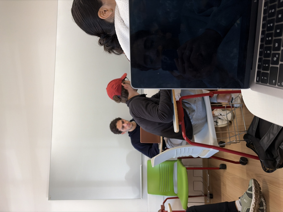 The image captures an indoor scene within a classroom or study area in Mexico City, Mexico, depicting multiple individuals seated and engaged with electronic devices. The perspective is from a seated position, looking forward and slightly to the left.
In the immediate foreground, a silver laptop, identified as a MacBook, rests on a white tabletop. Its dark, reflective screen displays the partial reflection of a person holding a dark smartphone, presumably the photographer. A black bag is positioned on the light-colored wooden floor beneath the table. To the far left foreground, a leg and foot, clad in a dark pant and a grey and white athletic shoe with a thick, textured sole, are visible.
In the mid-ground, a male individual is seated facing forward, wearing a red baseball cap, a dark long-sleeved shirt, and light grey sweatpants. He occupies a white plastic chair with a red metal frame and wheels, his head tilted downwards towards an obscured device. His feet, wearing white sneakers, are visible on the floor. To his left, a second male individual with dark, curly hair wears a dark blue long-sleeved top and is seated at a table, focused on a brown open laptop. An empty bright green plastic chair with a metal frame is positioned to the left of this second individual.
To the right of the central male individual, the back of a person's head and upper shoulder are visible. This individual has dark hair tied back in a bun and wears a light-colored, possibly white, fuzzy garment. A small gold earring is discernible on their right earlobe.
The background features a plain white wall, with a large, rectangular white board or panel mounted high. The floor is composed of light-colored wood. All visible individuals appear focused on their respective devices, with no direct interactions between them apparent.