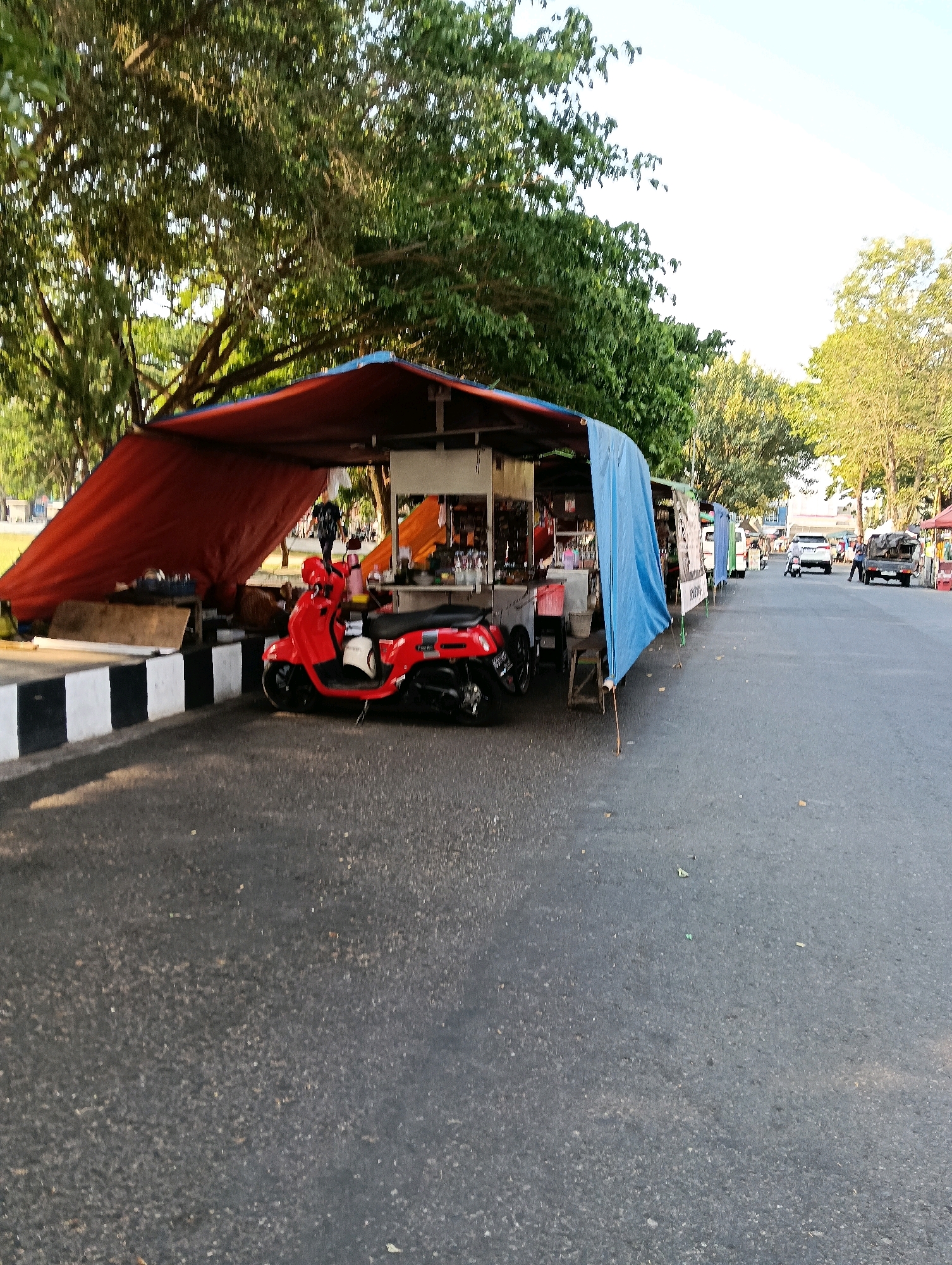 This is a photograph taken from a low angle, looking down a street lined with roadside stalls in Karanganyar, Indonesia. The stalls are covered with blue and red tarps, providing shade. A bright red scooter is parked in front of one of the stalls. The street is asphalt, and the sky is visible above the trees that line the area. Several cars and a truck are present on the street further down. The scene appears to be active with the presence of the vehicles and the stalls suggesting commerce. The lighting indicates it is likely late afternoon or early morning, given the long shadows cast.