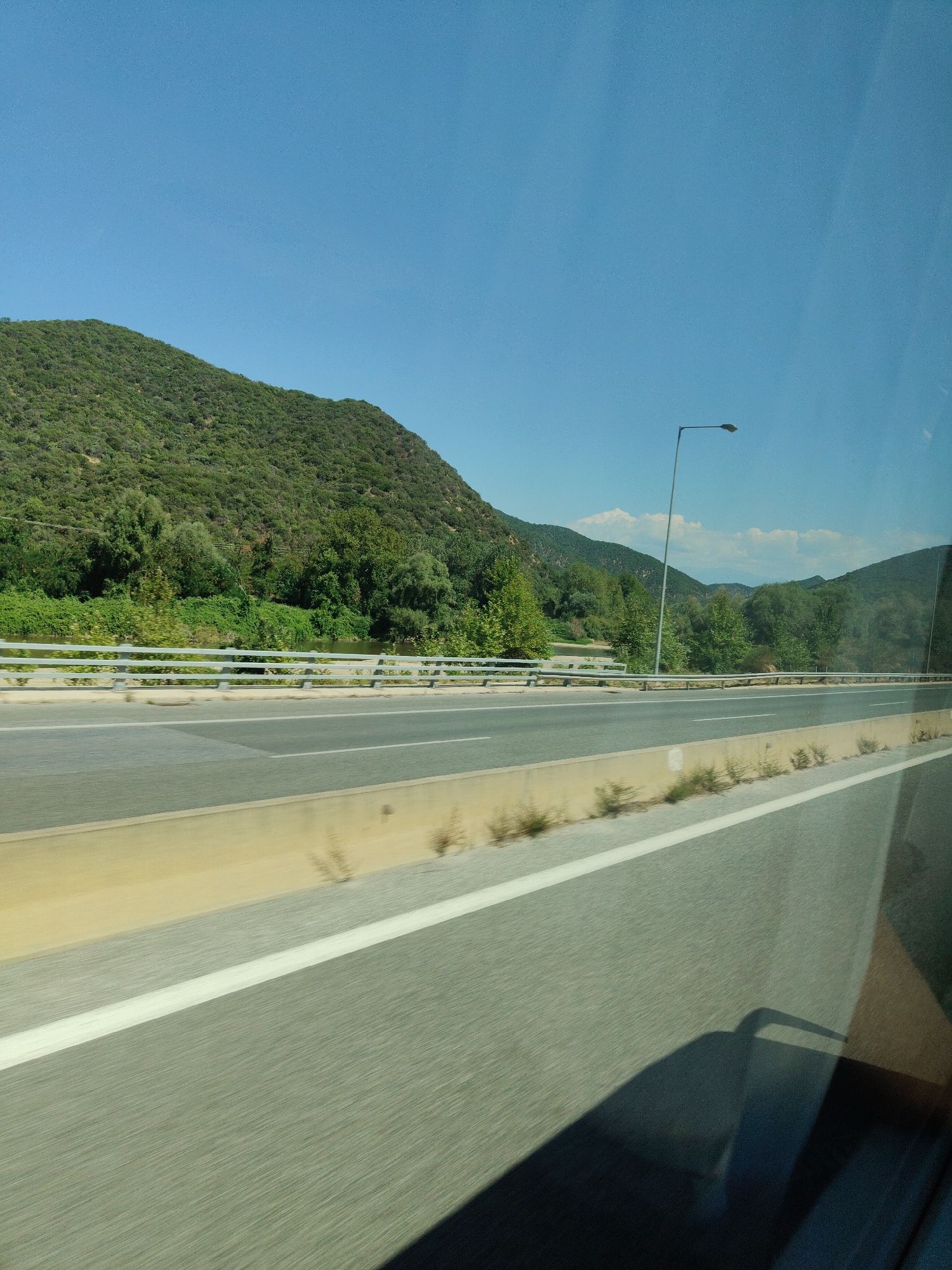 This image depicts a view from a vehicle traveling on a highway. The highway features two lanes of asphalt with white lane markings. A yellow barrier runs along the side of the road, bordered by sparse vegetation. In the background, a lush green hillside rises, covered in trees and foliage. A metal guardrail separates the highway from a body of water and more dense vegetation. A tall lamppost is visible on the right side of the highway, with the sky above being a clear, bright blue. The perspective suggests the photo was taken while moving, evidenced by the motion blur on the road. The location is identified as the Municipal Unit of Promachonas, Greece.