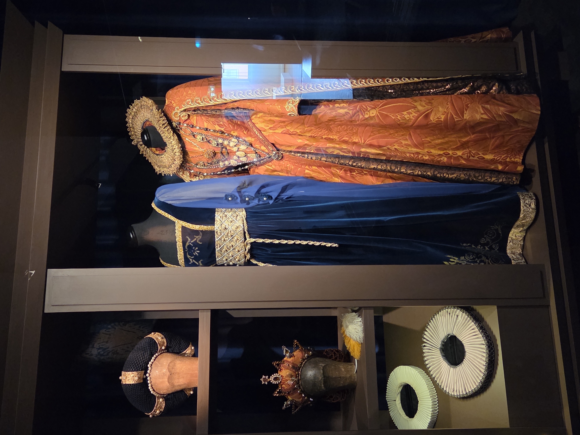 A dimly lit museum display features historical costumes and headwear within a dark wooden cabinet. On the top shelf, a black and gold headdress with pearls is displayed on a wooden head form. Below that, on the second shelf, a jeweled orange and brown crown sits atop another wooden head form, accompanied by a few feathers. The bottom shelf holds two white, pleated neck ruffs.
Dominating the right side of the display are two mannequins wearing elaborate historical dresses. The mannequin on the left wears a floor-length, deep blue velvet gown with gold trim around the neckline and a decorative gold belt. The mannequin on the right is dressed in a striking orange and gold brocade gown with intricate patterns, adorned with a large, ornate ruffled collar and multiple layered necklaces. The garments are positioned behind glass, reflecting a faint light from what appears to be a window or another display area in the background. This display is located at the Malmö Museums in Malmö, Sweden.