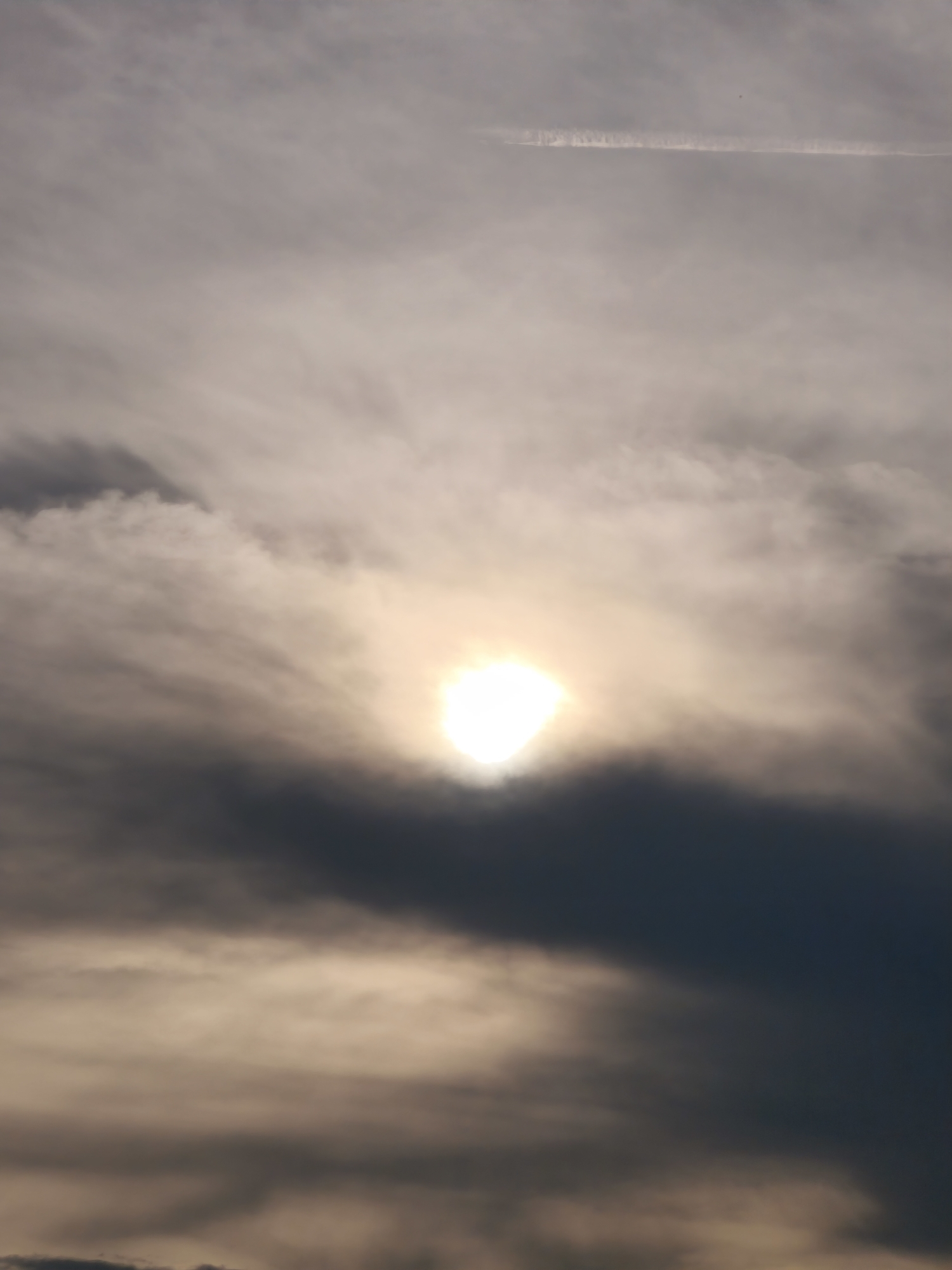 The image captures a segment of the sky above Passignano sul Trasimeno, Italy. Dark clouds dominate the lower half of the frame, with lighter, wispy clouds above them. The sun is a bright, luminous orb partially obscured by the clouds, creating a halo effect. A faint contrail from an aircraft is visible in the upper portion of the sky. The overall impression is one of a dramatic sky, possibly before or after a weather event. There are no discernible people, specific subjects, or actions depicted in the image. The environment is solely the sky.