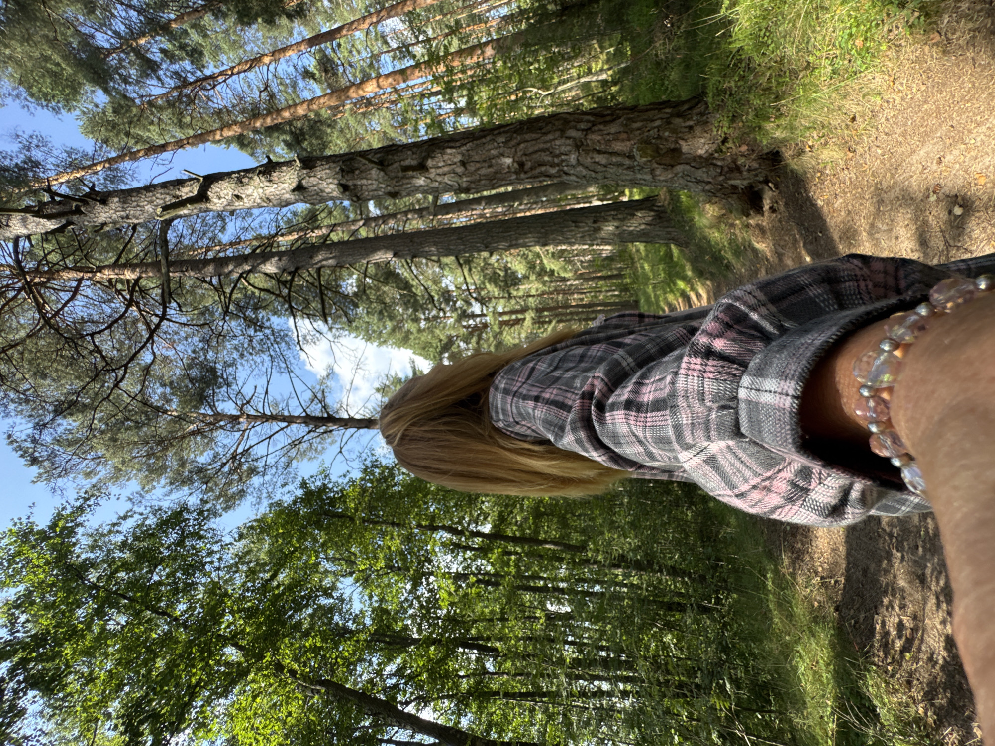A first-person perspective shot captures a person in a forest. The subject is wearing a plaid shirt with pink and gray colors and a bracelet on their wrist. The forest setting is characterized by tall pine trees, lush green foliage, and a path winding through the trees. Sunlight filters through the canopy, creating dappled light on the forest floor. The location is specified as Portovoe, Russia.