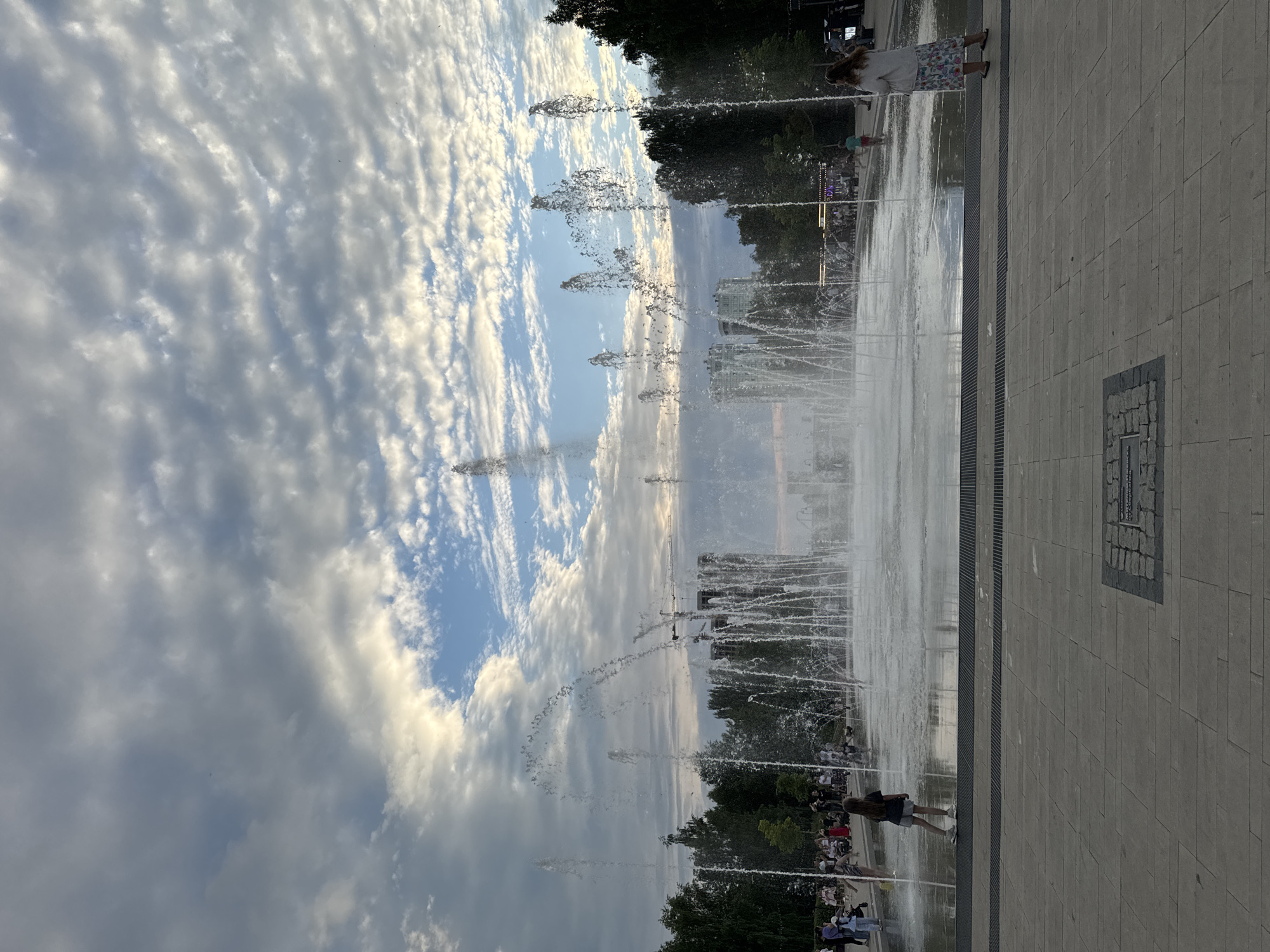 The image shows a wide shot of a public park with a large fountain. The fountain is the central focus, with water jets spraying into the air, creating a dynamic visual. People are scattered around the fountain, some walking, others standing and observing. The park appears to be well-maintained, with paved walkways and a grassy area with trees in the background. The sky is partly cloudy, with a mix of bright white clouds and patches of blue sky, suggesting it's daytime, possibly late afternoon or early evening given the soft light. The overall mood seems peaceful and relaxed, with people enjoying the public space. There are no visible signs or text in the image. The location is identified as Izhevsk, Russia.