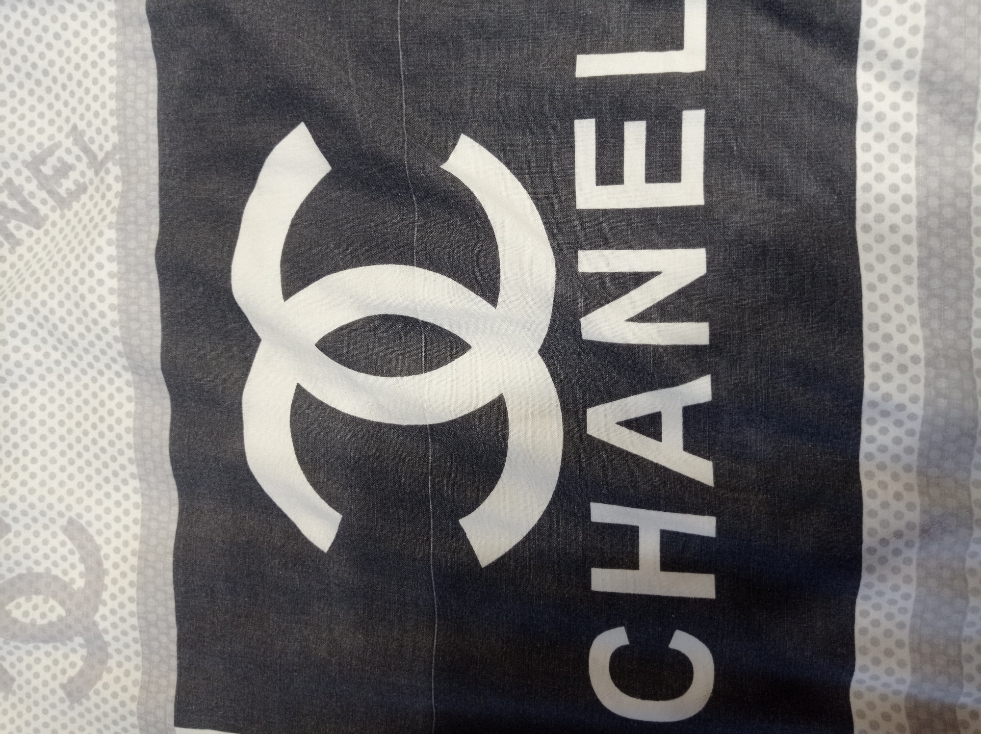 The image displays a close-up view of a black fabric with a prominent white Chanel logo and the word "CHANEL" written in white capital letters. The logo consists of two interlocking "C" shapes. Above the black fabric, there is a white section with a pattern of gray dots and a faint outline of the Chanel logo. Below the black fabric, there is a similar white section with a gray dotted pattern and horizontal gray stripes. The fabric appears to be part of a textile, possibly a scarf or bedding. There are no people, cityscapes, or specific environmental features visible that would indicate a location in Mukachevo, Ukraine. The image is a product shot or detail of the fabric itself.