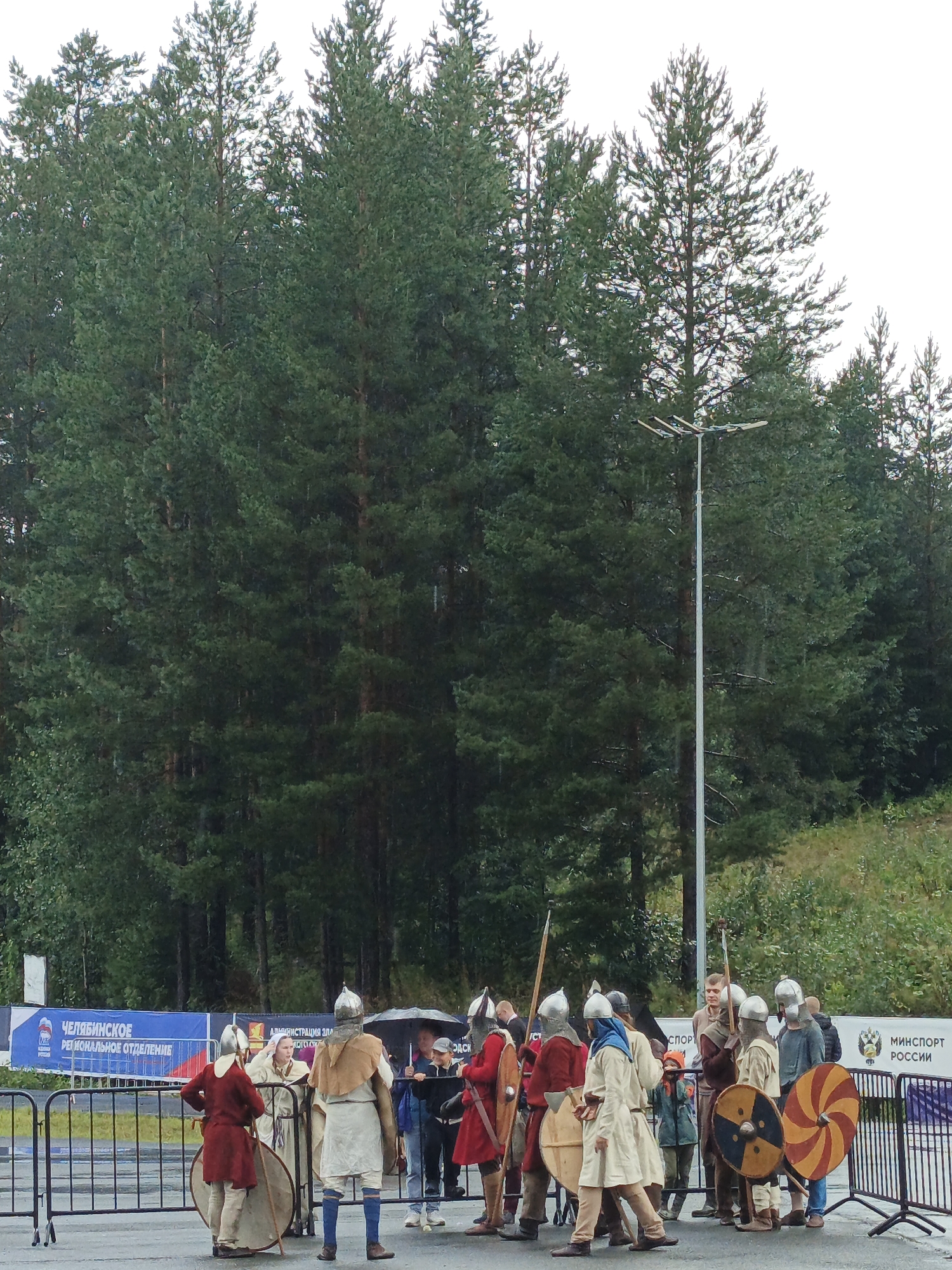 In Zlatoust, Russia, a group of individuals dressed in historical warrior attire, featuring helmets, tunics, and shields, are gathered outdoors. The scene appears to be a reenactment or historical event. Some individuals are holding spears and axes. In the background, a dense forest of tall evergreen trees dominates the landscape. A tall, modern lamp post stands amidst the trees. There is a blue banner in the background with Russian text, possibly indicating an event sponsor or organizer. Several people in modern clothing are also present, some observing the reenactment, one holding an umbrella, and others interacting with the participants. The ground is wet, suggesting recent rainfall.