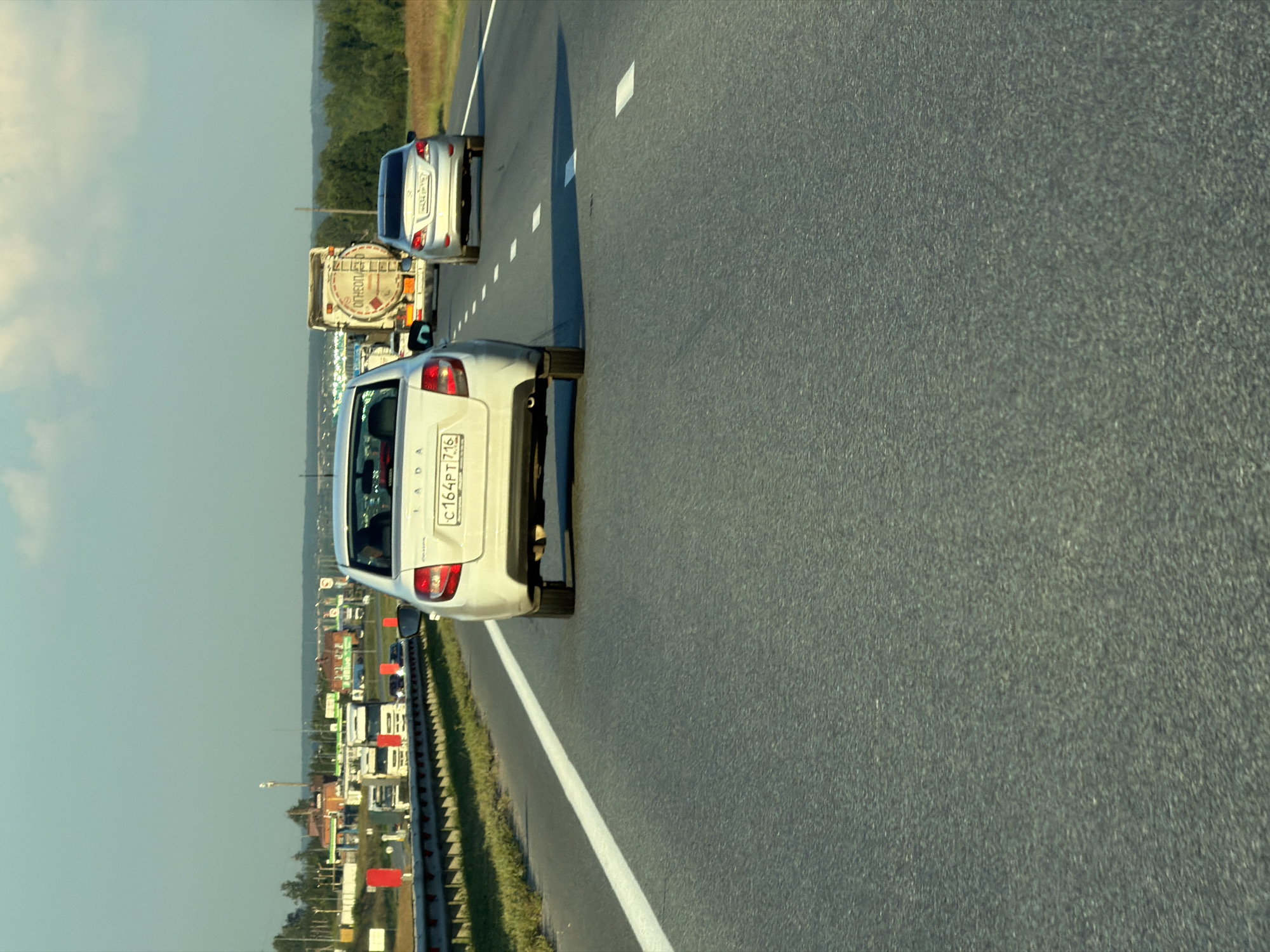 A white Lada Granta sedan travels on a multi-lane asphalt highway in Aryshkhazda, Russia. The car is in the right lane, with its rear facing the camera. A silver sedan is in the left lane, further ahead. A large tanker truck is visible in the distance, in the center lane. The highway is bordered by a metal guardrail and a grassy median. The sky is partly cloudy, and the sun is setting, casting long shadows. The scene appears to be set in a rural or semi-rural area, with trees and buildings visible in the background.