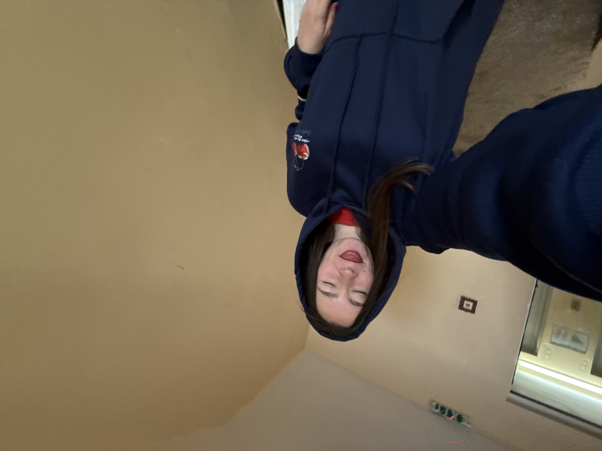 A medium shot depicts a woman indoors in Sofia, Bulgaria. She is wearing a blue hoodie and a red garment underneath, with the hood pulled over her head. She has long, dark hair. Her eyes are closed and she is sticking out her tongue slightly.
The background consists of a light tan wall. In the lower right corner, there is a window or a bright light source illuminating the area. Just above it are some electrical outlets and a small square object mounted on the wall. The photograph is slightly rotated counter-clockwise.