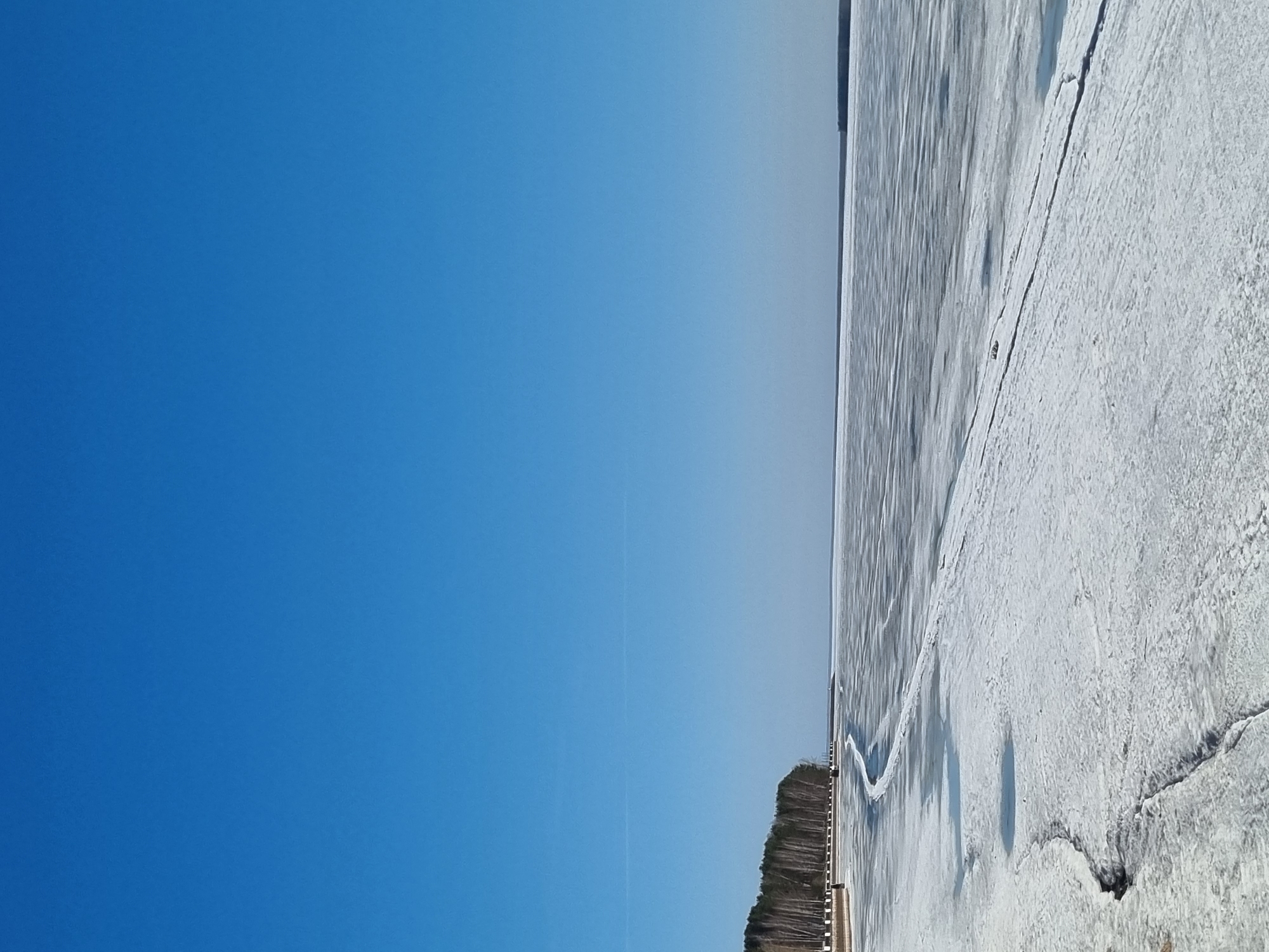 This image captures a serene winter or early spring landscape in Chaikovsky, Russia. The scene is dominated by a vast, brilliant blue sky that occupies the upper portion of the frame, appearing cloudless and vibrant. A faint, thin white line, possibly a contrail, stretches horizontally across the sky.
Below the expansive sky, a distant, dark green or brown band of coniferous trees marks the shoreline, forming a clear horizon line. The trees appear dense, characteristic of a cold-climate forest.
The lower half of the image is covered by a wide expanse of frozen water, likely a lake or large river. The ice surface is rugged and textured, with visible cracks and patches of snow. Some areas show clearer, darker ice, possibly revealing refrozen water, creating abstract patterns and gentle undulations across the surface. The overall impression is one of a stark, frozen wilderness.
No individuals or specific activities are visible, contributing to the tranquil and still nature of the landscape. The bright, clear sky and strong lighting suggest it is midday or early afternoon. The presence of extensive ice confirms cold, sunny weather conditions typical of winter or early spring in this region. There is no visible text within the image.