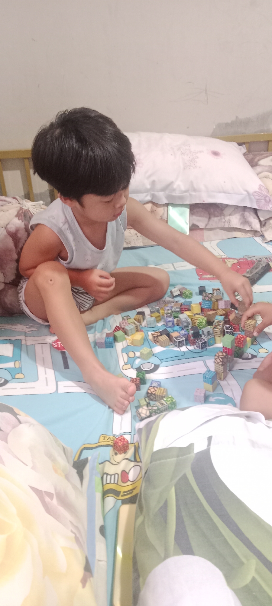 A young child is sitting on a bed, playing with a collection of small, colorful blocks. The blocks appear to be made of wood or plastic and are arranged in various configurations on the light blue bedsheet, which features a cartoonish road pattern with cars. The child, with dark hair and wearing a light blue tank top and striped shorts, is focused on manipulating the blocks with their right hand. Another person's hand, partially visible on the right side of the image, seems to be interacting with the blocks as well. The background shows a white wall and pillows on the bed. The overall scene suggests a casual indoor activity. This image was taken in Banyuraden, Indonesia.