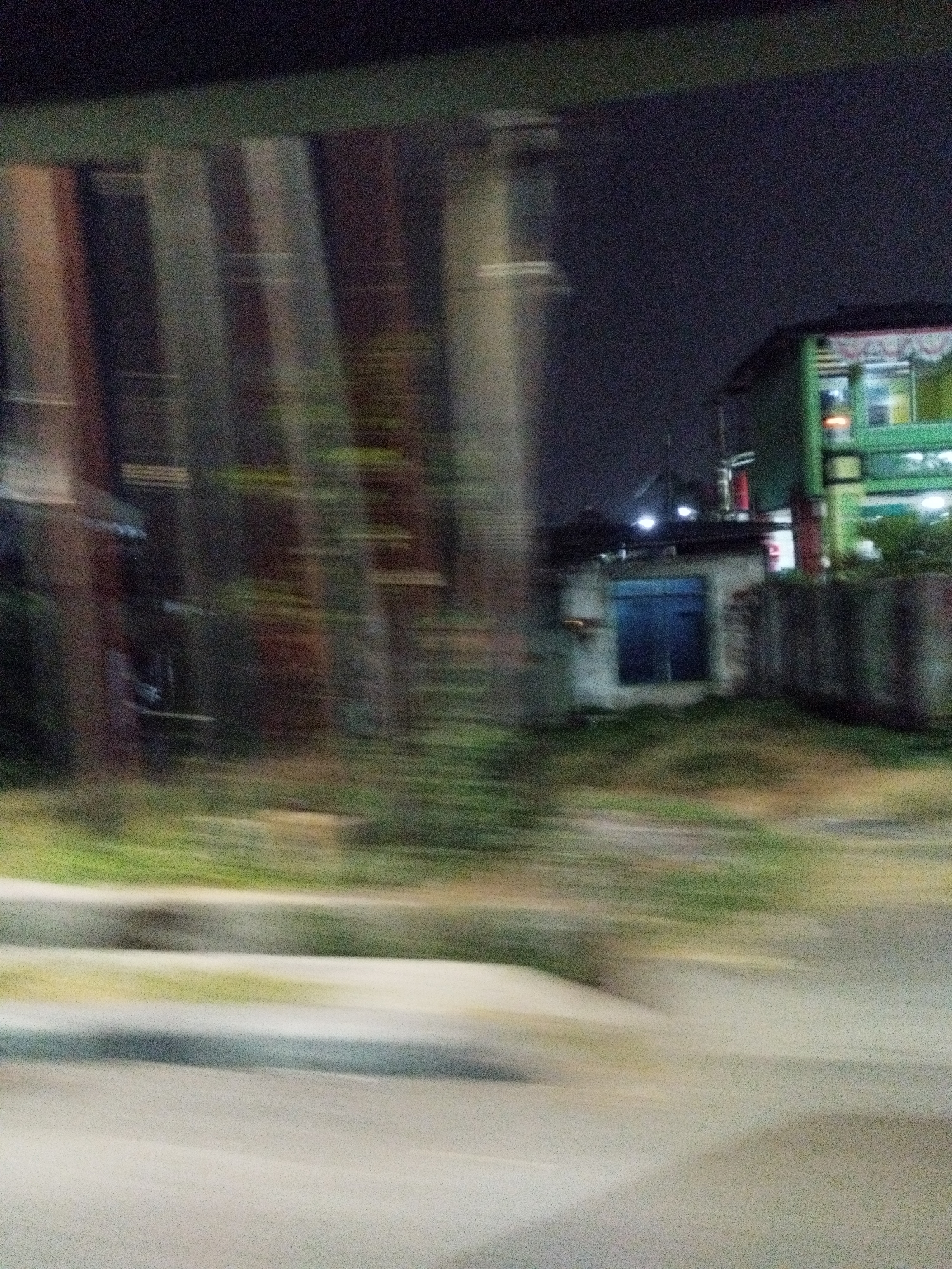The image is a motion-blurred photograph taken at night in an urban environment, likely along a road in Surabaya, Indonesia. The blur suggests movement, possibly from a vehicle. On the right side of the frame, a multi-story building with a green facade and illuminated windows is visible. To its left, a smaller, single-story structure with a blue door is partially obscured. In the foreground, a road surface and a concrete barrier are discernible. The background is dark with some indistinct shapes that could be trees or other urban structures. The overall scene depicts a typical nighttime street view in a developed area.