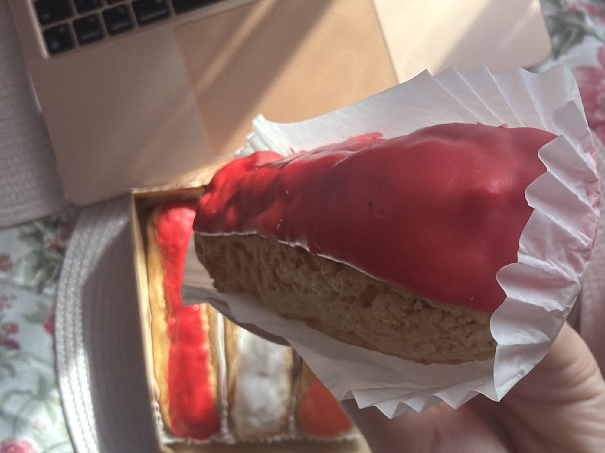 The image depicts a red-glazed eclair held in a white paper liner, with a box of eclairs visible in the background, positioned next to a laptop. The scene is situated on a floral tablecloth. The location is suggested to be Saint Petersburg, Russia, although there is no definitive visual evidence in the image to confirm this.