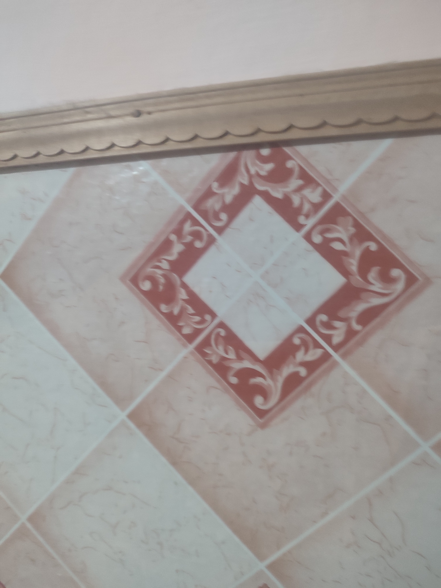 A decorative tile border runs along the top of an image showing a tiled wall. The tiles are square and laid diagonally, with a patterned diamond in the center of each. The colors are muted, with shades of pink, beige, and white. The border above is a light wood color with a scalloped edge. The scene appears to be indoors, likely in a bathroom or kitchen, given the tiling. There are no people or discernible actions visible. The lighting is soft and diffused. The image is slightly out of focus, giving it a soft, almost dreamlike quality. The location of this image is not specified as Chernivtsi, Ukraine.