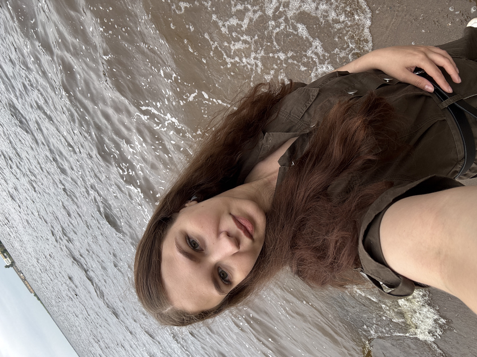 This is a vertically oriented, close-up selfie taken outdoors on a beach. The primary subject is a young woman with long, dark brown hair, fair skin, and light green eyes. She is wearing a dark green, collared shirt with pockets. She is looking directly at the camera, with a neutral or slightly smirking expression. Her left hand is resting on her chest, near her collarbone.
The setting is a beach, with water lapping at the shore. The water is choppy and appears to be a muted gray-brown color. There is foam from the waves on the wet sand. In the background, a small section of a distant shoreline with buildings is visible on the left side of the image. The sky appears overcast.
The image suggests a casual outing by the water. The lighting indicates it is daytime, but the overcast sky suggests it might be a cloudy day. The location is identified as Chaikovsky, Russia. There is no visible text in the image.