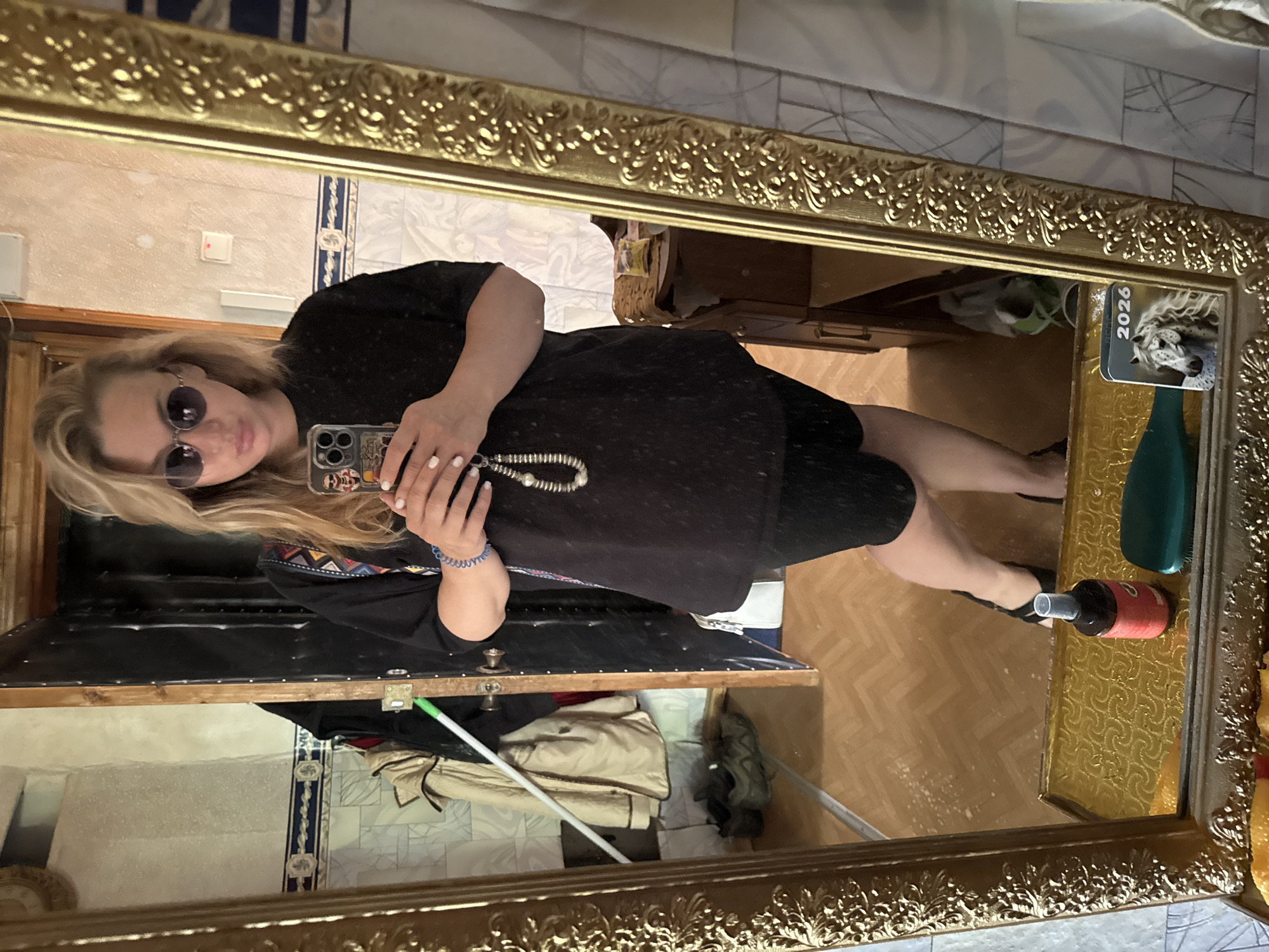 A woman with blonde hair and sunglasses takes a mirror selfie. She is wearing a black t-shirt, black shorts, and black sandals. She has a beaded necklace and a bracelet on her left wrist. She is holding a phone in her right hand. In the background, there is a door, a coat rack with jackets, and a mirror on the floor. The image appears to be taken in a residential setting, possibly an apartment in Valday, Russia.