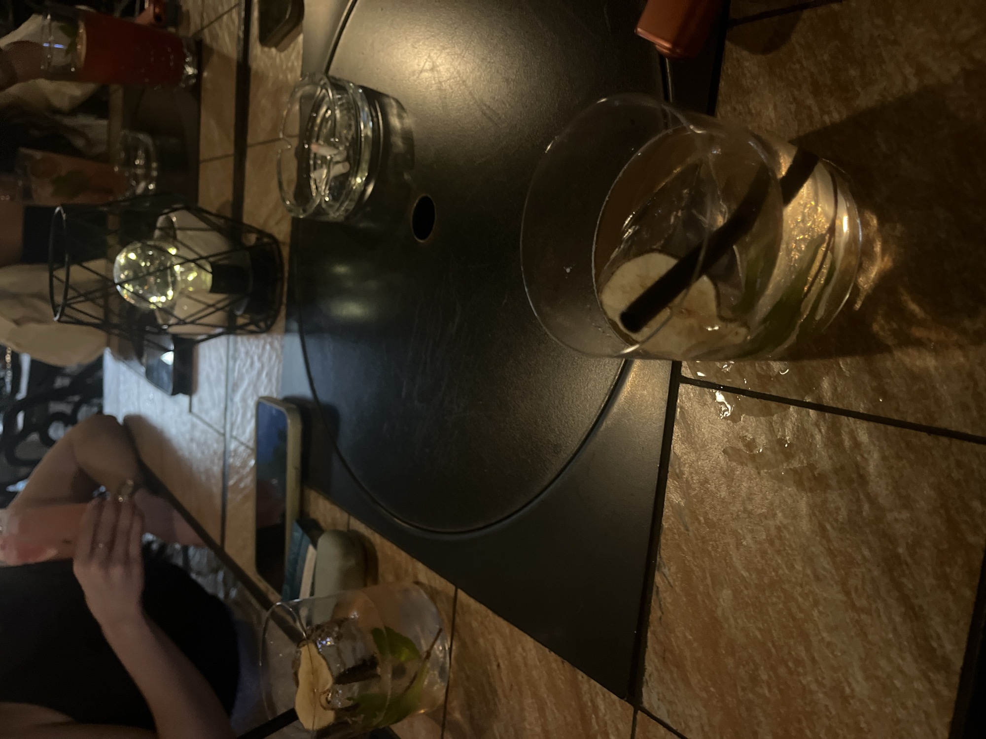 A group of individuals are gathered at an outdoor seating area at night. The scene is dimly lit, with the primary light source appearing to be a decorative lantern with a lit bulb. On a table, several drinks are visible, including a pink-colored beverage in a tall glass, a red-colored drink in a glass, and two other drinks in shorter glasses with ice and garnishes. An ashtray with cigarette butts is also on the table. A mobile phone is placed on the table near a stack of what appears to be coasters. One individual is partially visible, wearing a black tank top and holding a glass. The table itself is round and appears to be made of a dark material, possibly metal. The surrounding area has tiled flooring and appears to be part of a restaurant or bar setting.
