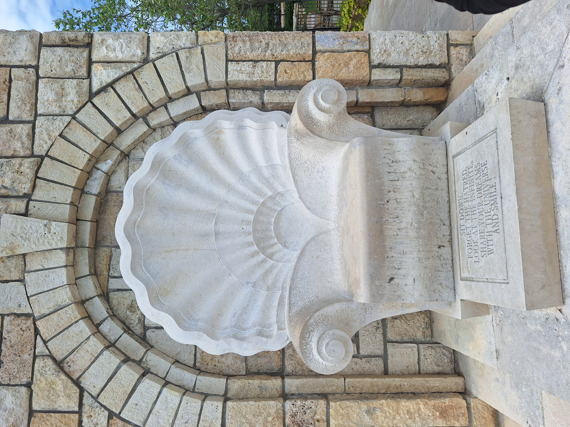 The image shows a stone structure, likely a seat or decorative element, set into a curved stone wall. The main structure features a large, stylized seashell shape, carved from white stone. To the right of the shell, a vertical rectangular stone tablet displays carved text. The text reads "SIT DOWN. FORGET THE HATRED, LOOK AT THE INFINITE, SHARE YOUR DREAMS WITH THE UNIVERSE AND SMILE!". The stone wall behind and around the structure is constructed of light-colored rectangular blocks. There is a tree visible above the wall, and the sky is partially visible in the background. This structure is located in the city of Varna, Bulgaria.