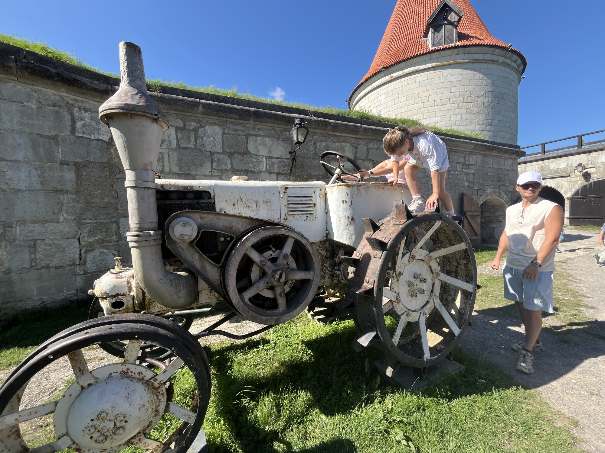 A young girl with blonde hair pulled back in a ponytail is climbing on an old, rusted white tractor. She is wearing a white t-shirt with a colorful design on the front and light pink shorts. Her left hand is on the steering wheel, and she is looking down at the tractor's controls. Next to the tractor, a man wearing a white sleeveless t-shirt, blue and white striped shorts, and a white baseball cap is standing and looking towards the tractor. He is also wearing glasses. The tractor is positioned on a grassy area in front of a stone wall with a green ledge on top. In the background, a circular stone tower with an orange roof is visible. The sky is clear and blue. This scene is located at Kuressaare Castle in Kuressaare, Estonia.