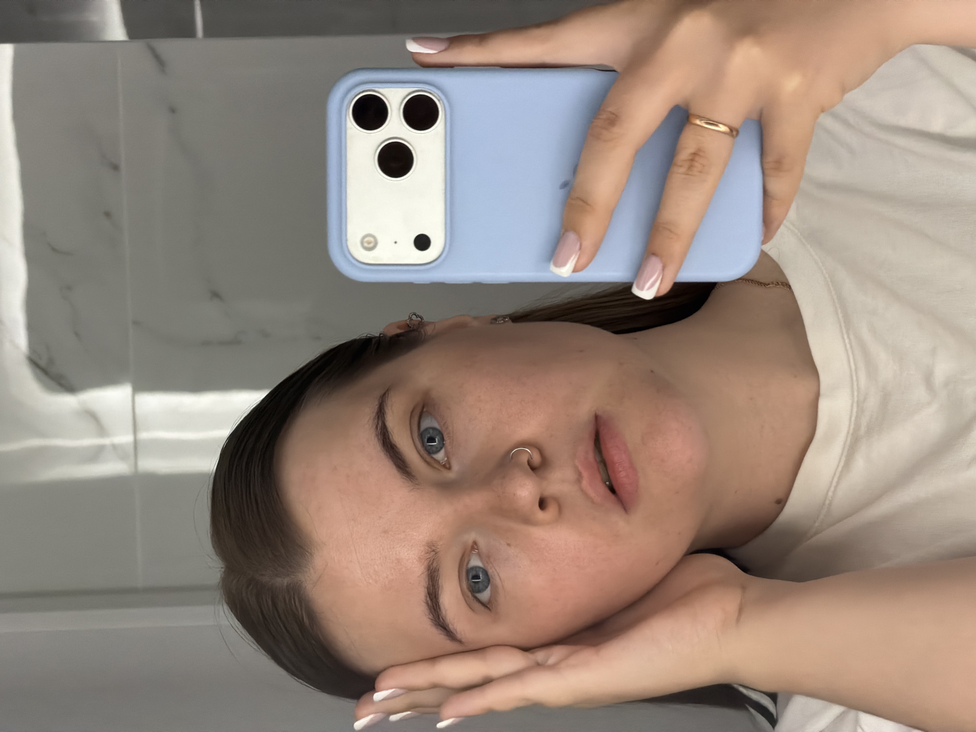 A young woman is holding a blue smartphone up to take a selfie. The woman has fair skin, blue eyes, and dark brown hair. She is wearing a white t-shirt. She is wearing a gold ring on her left hand and a nose ring. The background appears to be a bathroom with white marble-like tiles. The location is the city of Chaikovsky, Russia.