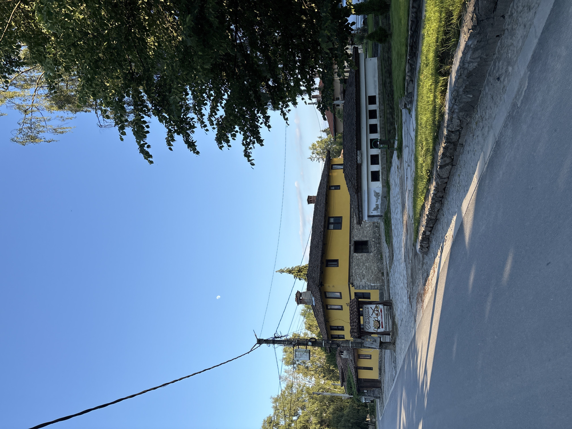 The image shows a street scene in Koprivshtitsa, Bulgaria, on a clear day with a bright blue sky. A road with shadows stretches from the foreground towards the left. On the right side of the road, a traditional yellow building with a stone foundation and a tiled roof stands prominently. Adjacent to it is a lower, white building with multiple square windows. A power line crosses the sky from the left to the right, supported by a utility pole. Trees line the background. A sign with Bulgarian text and an image of food is visible near the utility pole. The moon is faintly visible in the sky. A low stone wall with grass borders the sidewalk in front of the buildings.