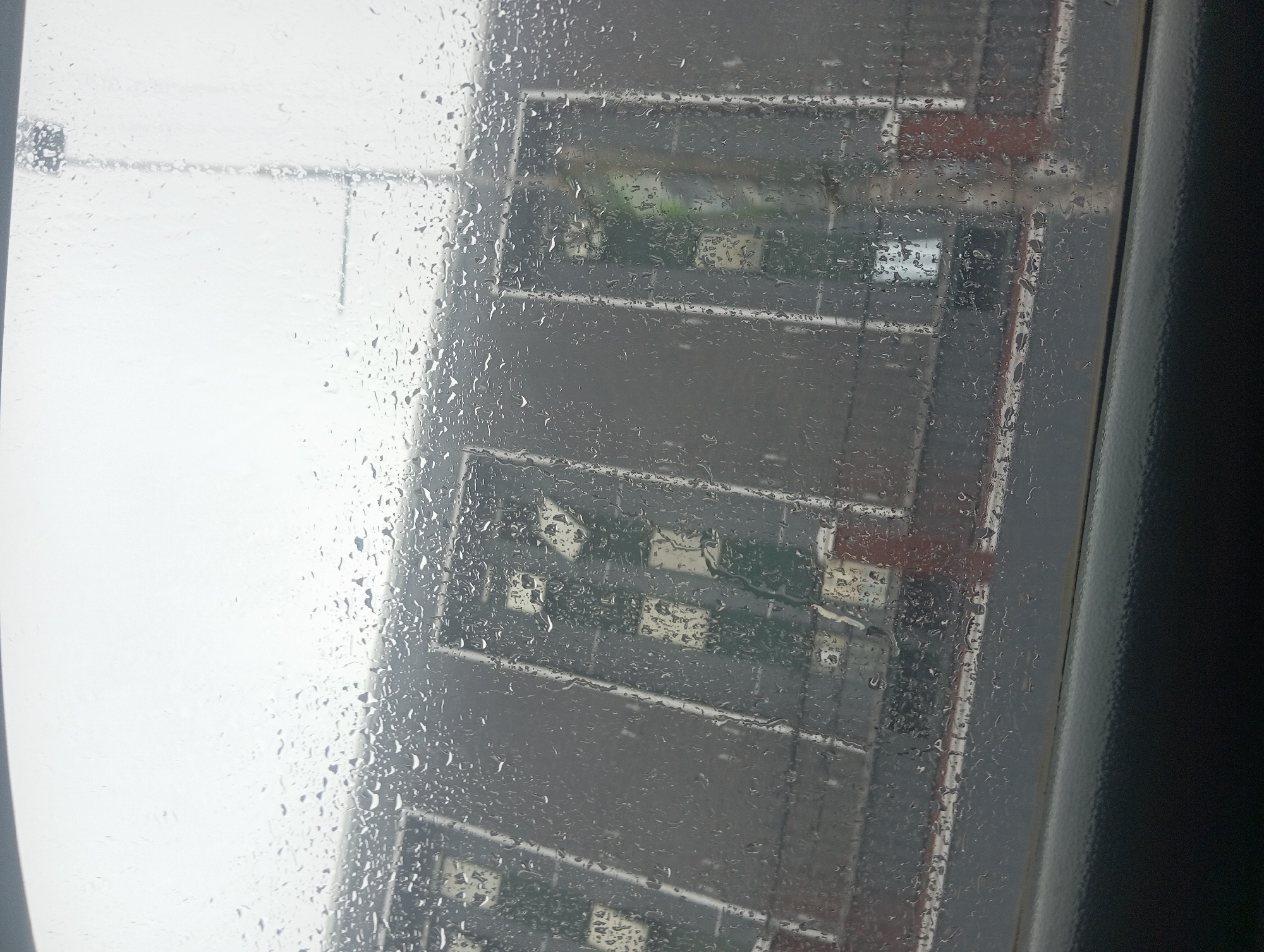A rainy day view from inside a vehicle in Onitsha, Nigeria, shows a row of buildings with distinctive light green and cream colored panels, possibly indicating a market or commercial structure. Water droplets obscure the view, distorting the building's facade and creating a blurry effect. The sky is overcast, contributing to the dull, muted color palette of the scene.