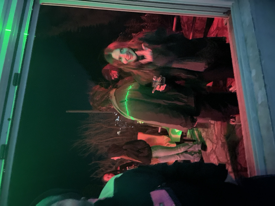 A group of people are visible through a doorway, lit with green and red hues. A woman with long hair smiles at the camera, while another person holds a drink. Several other figures are present, partially obscured. The scene appears to be an outdoor gathering at night. A bare tree is silhouetted in the background. The composition is vertically oriented.