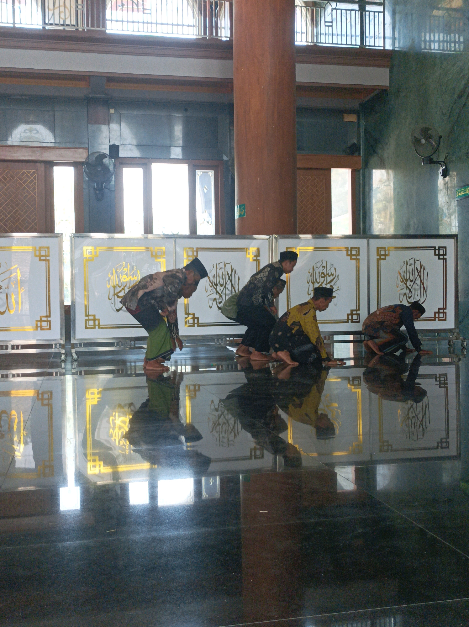Four men in traditional Islamic attire are performing prayer on the polished floor of a mosque. The men are wearing sarongs and caps, and their attire includes patterned shirts and tunics. They are positioned in a line, facing away from the camera, and appear to be in the process of bowing or prostrating. The floor reflects the figures and the environment, creating a symmetrical visual effect. Behind them, a decorative wall with Islamic calligraphy is visible. The background shows architectural elements of the mosque, including large windows and columns. The scene is illuminated by natural light streaming in from the windows, casting shadows and highlights on the floor. This scene is located in the mosque of Rengel, Indonesia.