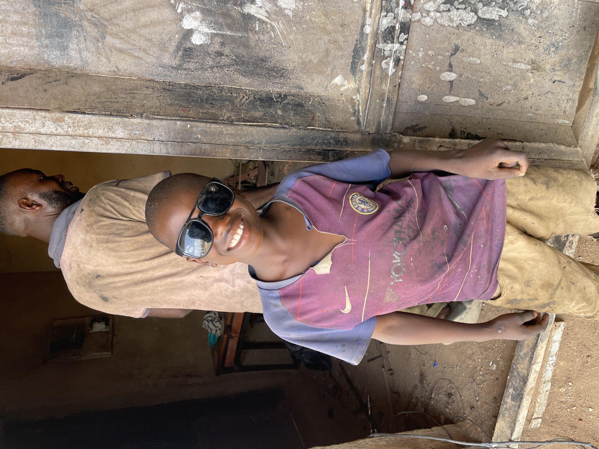 A young child stands smiling, wearing sunglasses and a tattered soccer jersey. An adult stands behind the child, partially visible. Both are in front of a metal door or gate that is partially open. The ground is dirt. The scene is likely located in Barakin Akawo, Nigeria, based on the user's request.
