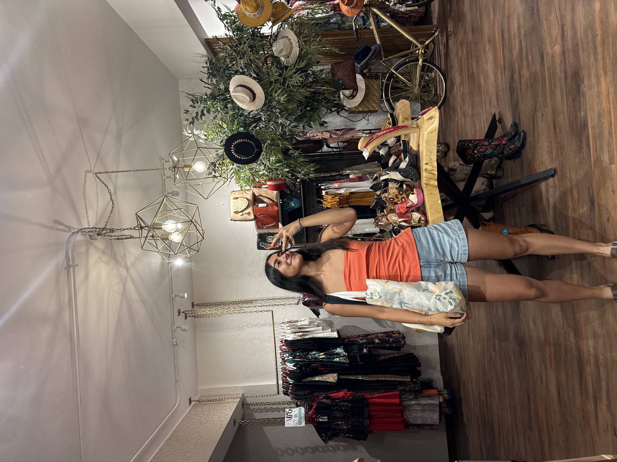 The image shows the interior of a clothing store. A woman in an orange top and denim shorts is holding a phone and appears to be taking a selfie. Racks of clothing are behind her. A display with hats, plants, a bicycle, and boots is located on the right. The floor is wood. Geometric light fixtures are hanging from the ceiling. The location is a clothing store in Domingo Arenas, Mexico.