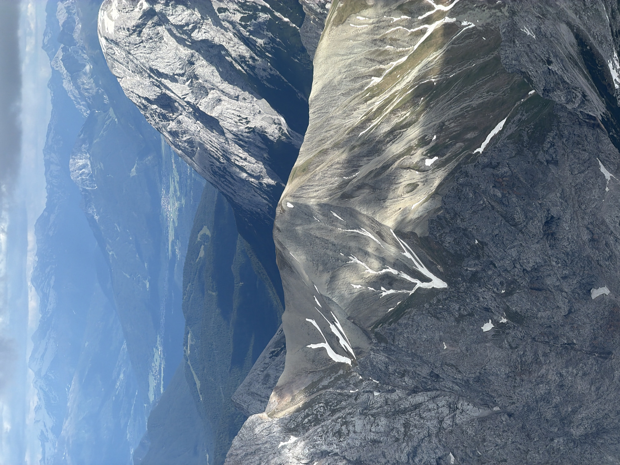 The image is an aerial view of the Zugspitze, Germany's highest peak, located near Garmisch-Partenkirchen. The photograph showcases a rugged mountain landscape with steep, rocky slopes and patches of snow. The foreground is dominated by the textured grey rock faces of the mountain, with some white snow streaks accentuating the contours. In the mid-ground, another mountain ridge, lighter in color, slopes downwards, also featuring sparse snow. Further in the distance, a valley is visible with a settlement, characterized by small buildings and green fields, nestled between dark green forested slopes. Beyond the valley, layers of progressively bluer, hazy mountains recede into the horizon under a cloudy sky. The scene depicts a vast, natural alpine environment. There are no people or objects indicative of human activity other than the distant settlement.
