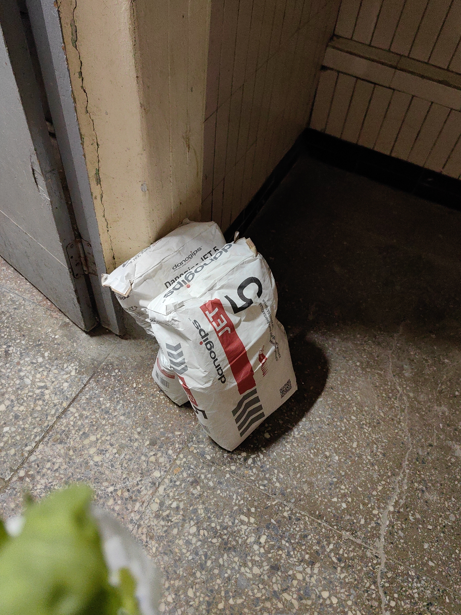Two white bags of building material, branded "danogips JET 5," are placed on a speckled concrete floor next to a gray door with visible hinges. The bags are positioned near a light yellow, textured wall with some visible cracks. The scene appears to be an indoor entryway or hallway, possibly in an apartment building in Minsk, Belarus.