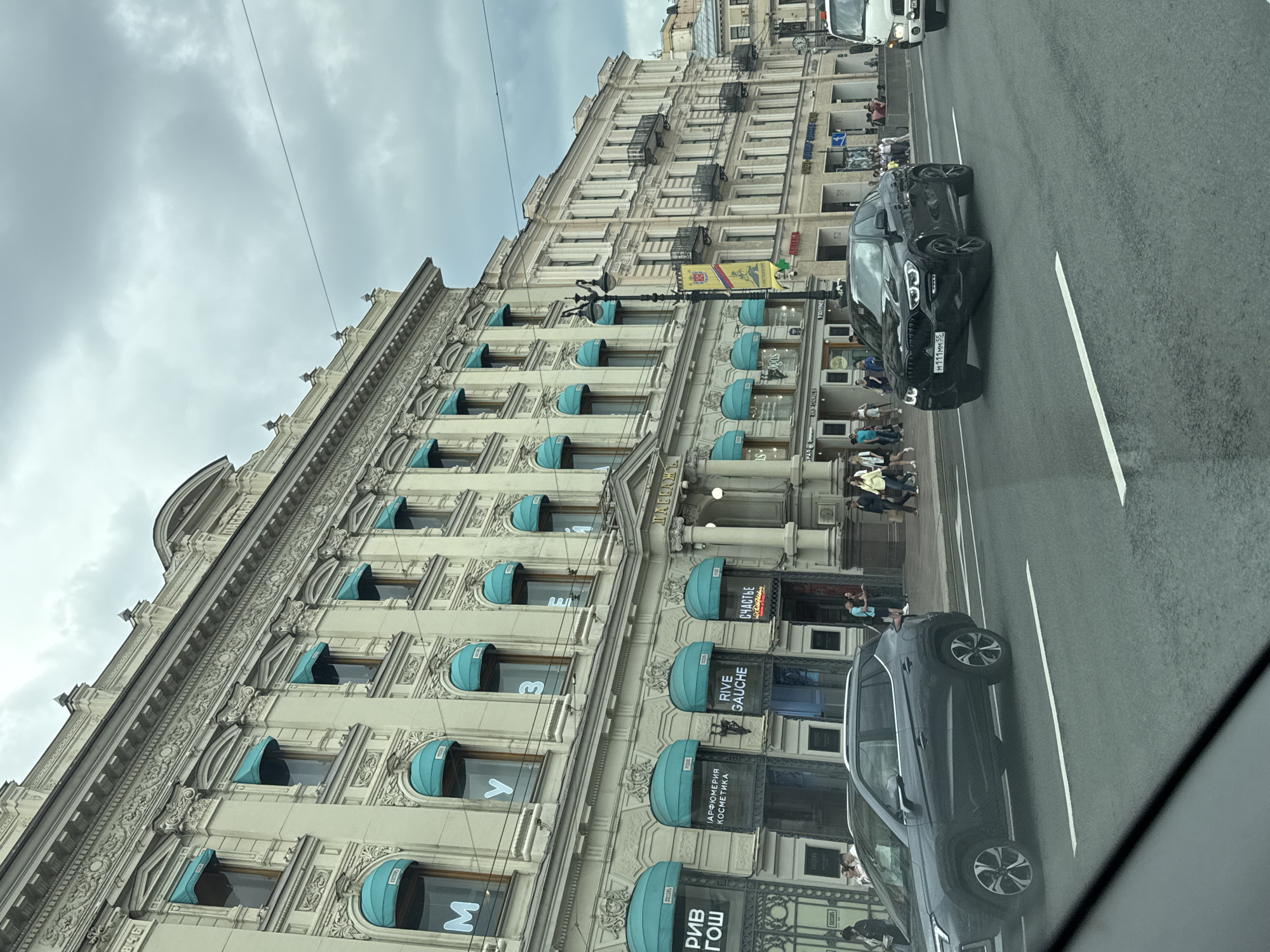 This image depicts a street scene in Saint Petersburg, Russia, likely on Nevsky Prospekt, given the architecture and the presence of prominent businesses. The focal point is a grand, ornate building with a light-colored facade, characterized by numerous windows adorned with teal awnings. The building features intricate architectural details, including decorative moldings and columns. Several storefronts are visible at the ground level, with illuminated signs indicating businesses such as "RIVE GAUCHE" and "СПОРТ ГОШ".
The street in the foreground is a multi-lane road with white lane markings. Two cars are captured in motion: a dark gray SUV on the left and a black BMW coupe on the right. Pedestrians are seen walking on the sidewalk alongside the buildings. The sky is overcast with clouds, suggesting a temperate weather day. The overall scene conveys a sense of urban activity and architectural grandeur.