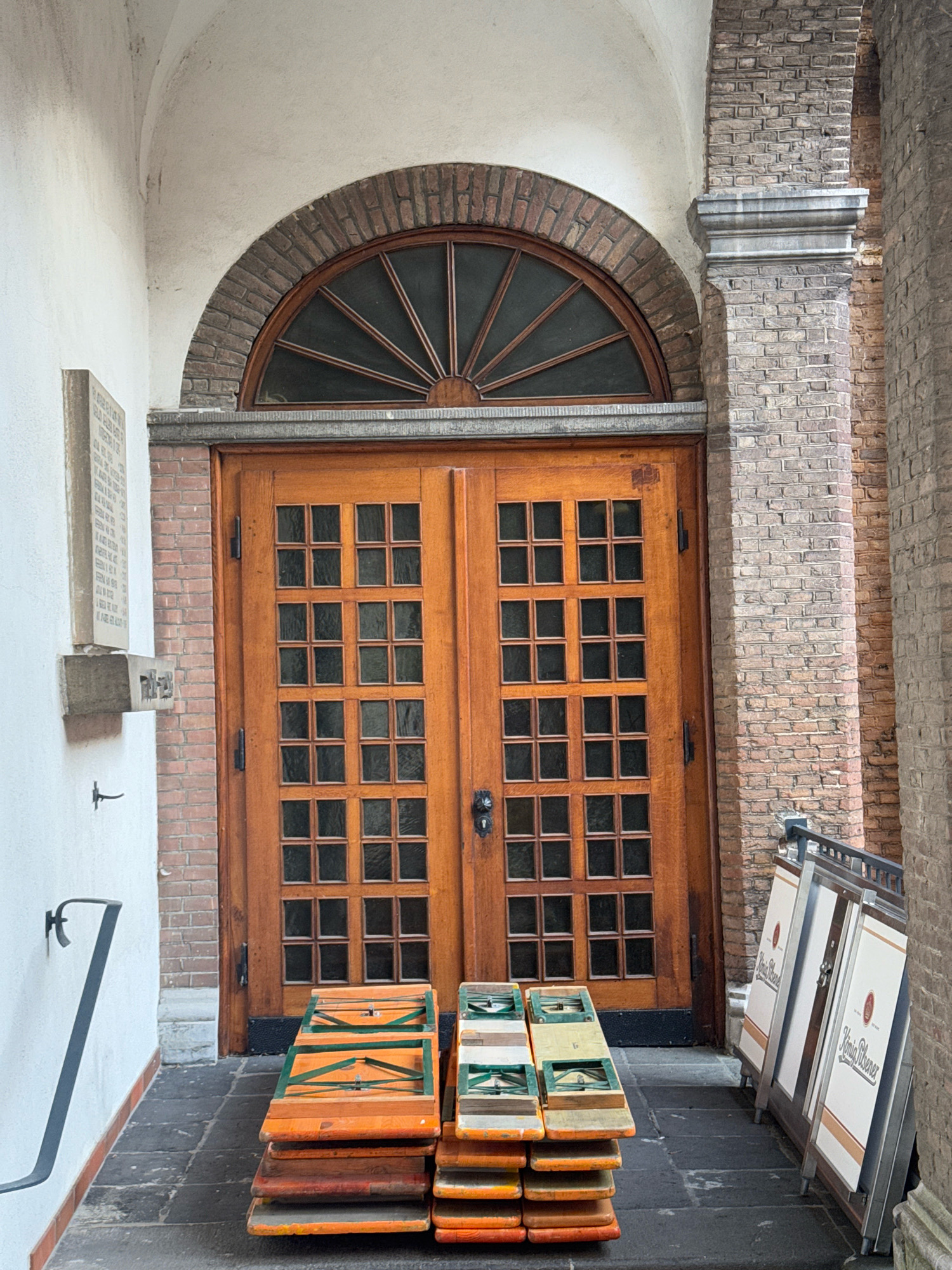 The image depicts an outdoor view of a building entrance in Cleves, Germany. The entrance features a large, double wooden door with many glass panes, framed by a brick archway and a stone pillar. To the left of the door, a white wall displays a stone plaque. In front of the door, several stacked wooden tables and benches are visible. To the right of the door, a row of A-frame advertising signs, some bearing the "König Pilsener" logo, are leaning against a brick wall. The ground is paved with dark flagstones. There are no people present in the image. The overall scene suggests a setting that could be a beer garden or an establishment that uses outdoor seating.