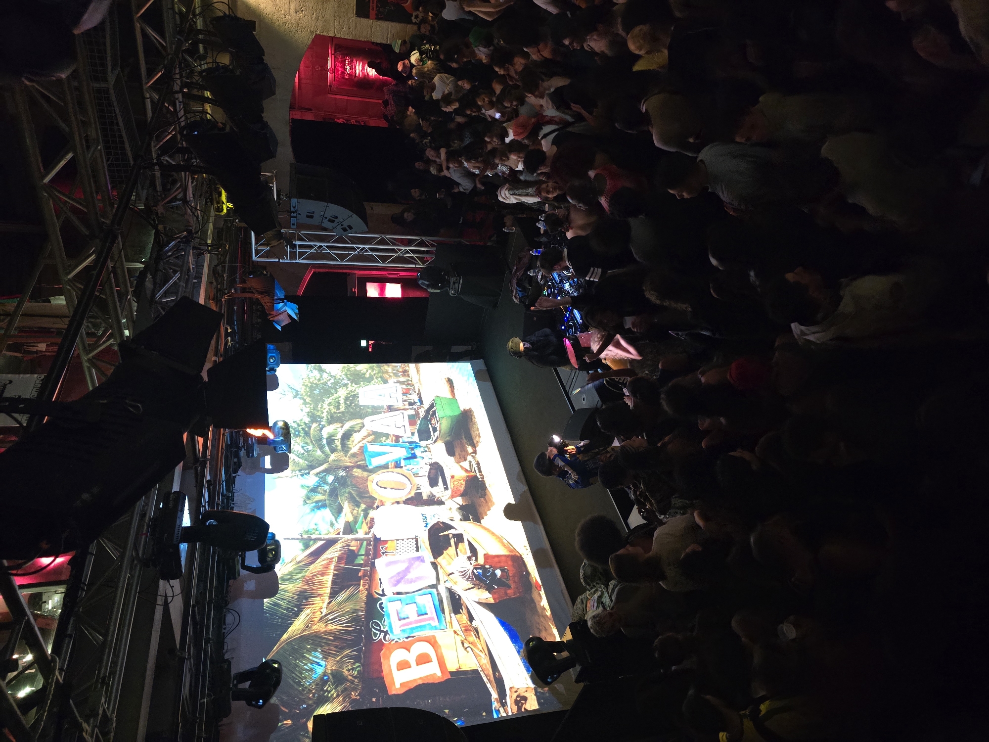 A nighttime concert scene in Paris, France, features a large crowd gathered in a confined space, likely a venue. Stage lighting equipment is visible on the left. A large screen displays an image with the words "BENOVAYA." A DJ booth is positioned on the ground, with the crowd closely surrounding the DJ. The environment has red architectural elements.