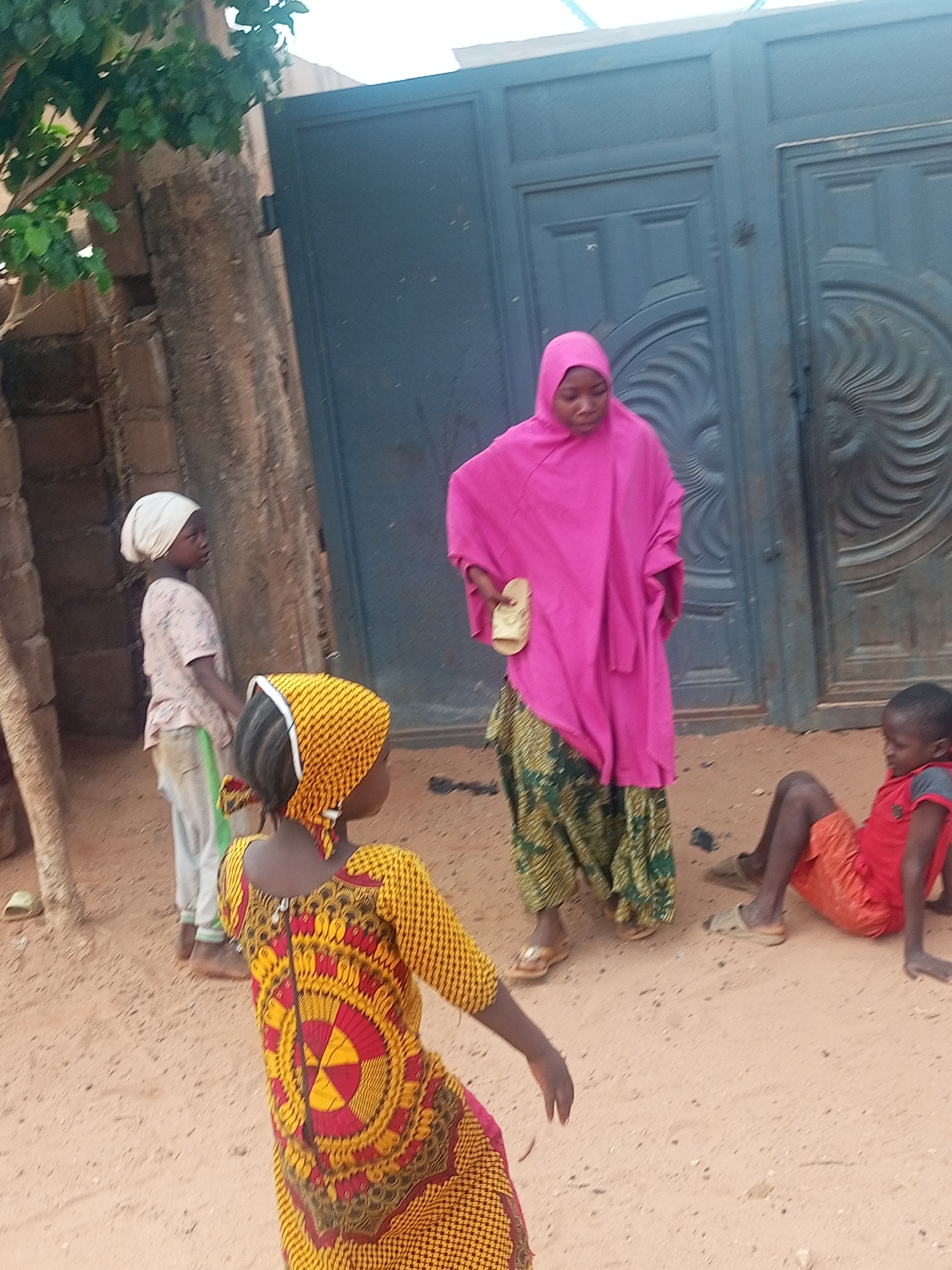 The image shows four people in an outdoor setting, likely a yard or open space in Tempure, Nigeria.
A woman wearing a pink headscarf and robe, and a long patterned skirt, is standing and holding a sandal in her hand.
Two girls are present. One is wearing a yellow and red patterned dress and headscarf and stands with her back to the viewer. The second girl, wearing a white headscarf and a light-colored shirt, stands in the background facing left.
A boy in a red shirt and orange shorts is sitting on the ground.
A gray brick wall is partially visible behind the girl wearing the white headscarf. A tall, dark-colored gate is visible behind the woman wearing pink. The ground appears to be sandy or dirt. A tree is visible on the left side of the image.