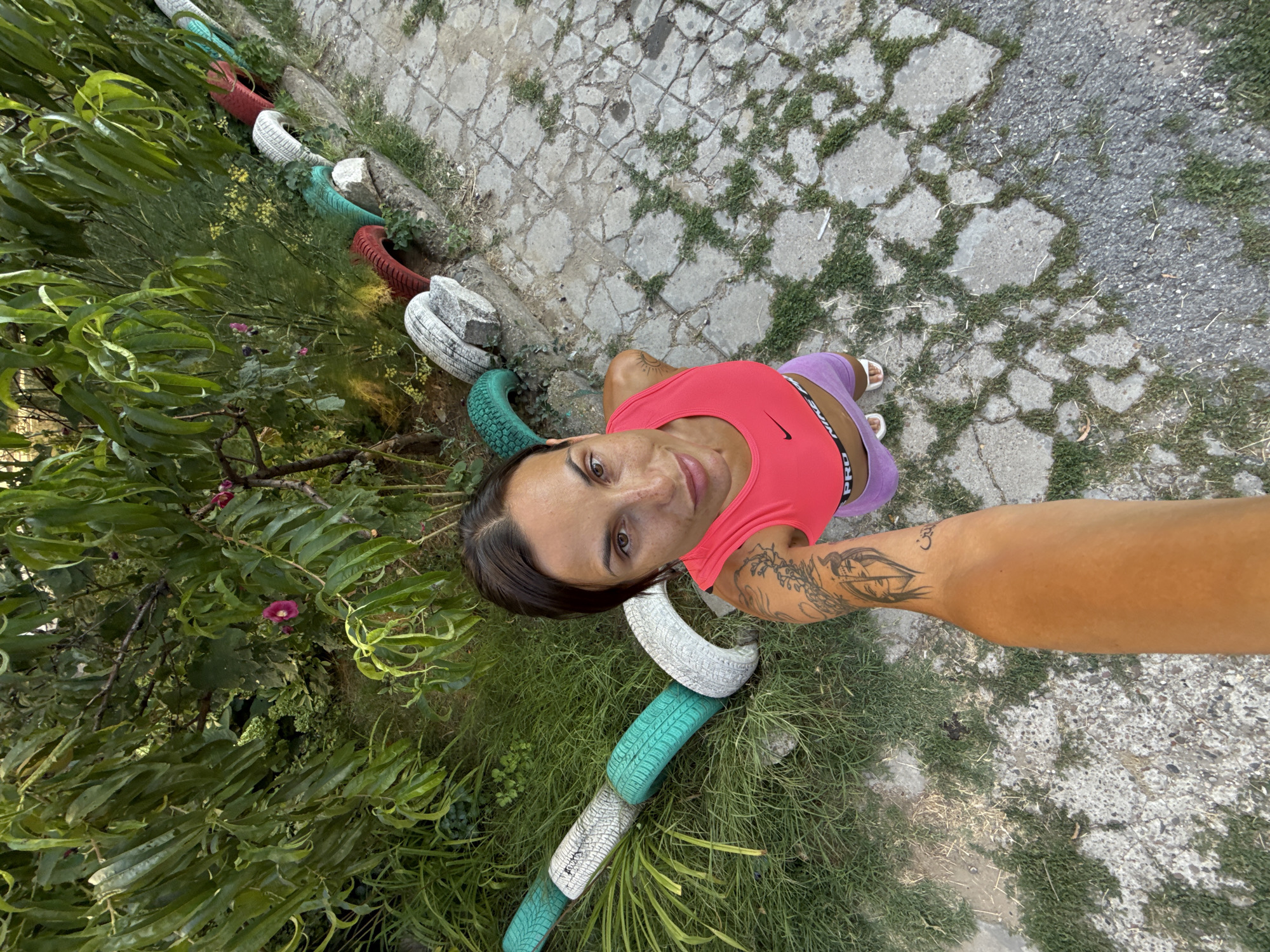 In Pleven, Bulgaria, a woman with dark hair, wearing a pink Nike sports bra and purple shorts, takes a selfie. Her left arm, adorned with tattoos, is extended towards the camera. She stands on a cracked concrete pathway with patches of green grass and dry foliage. Behind her, a garden bed is bordered by a row of colorful painted car tires – white, green, and red – and large grey stones. Lush green foliage and a small pink flower are visible in the background.